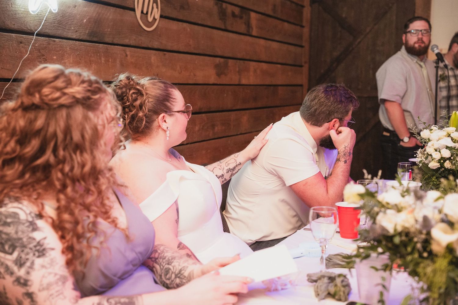 Emotional wedding reception moment with bride and groom at head table during heartfelt speech in rustic barn venue.
