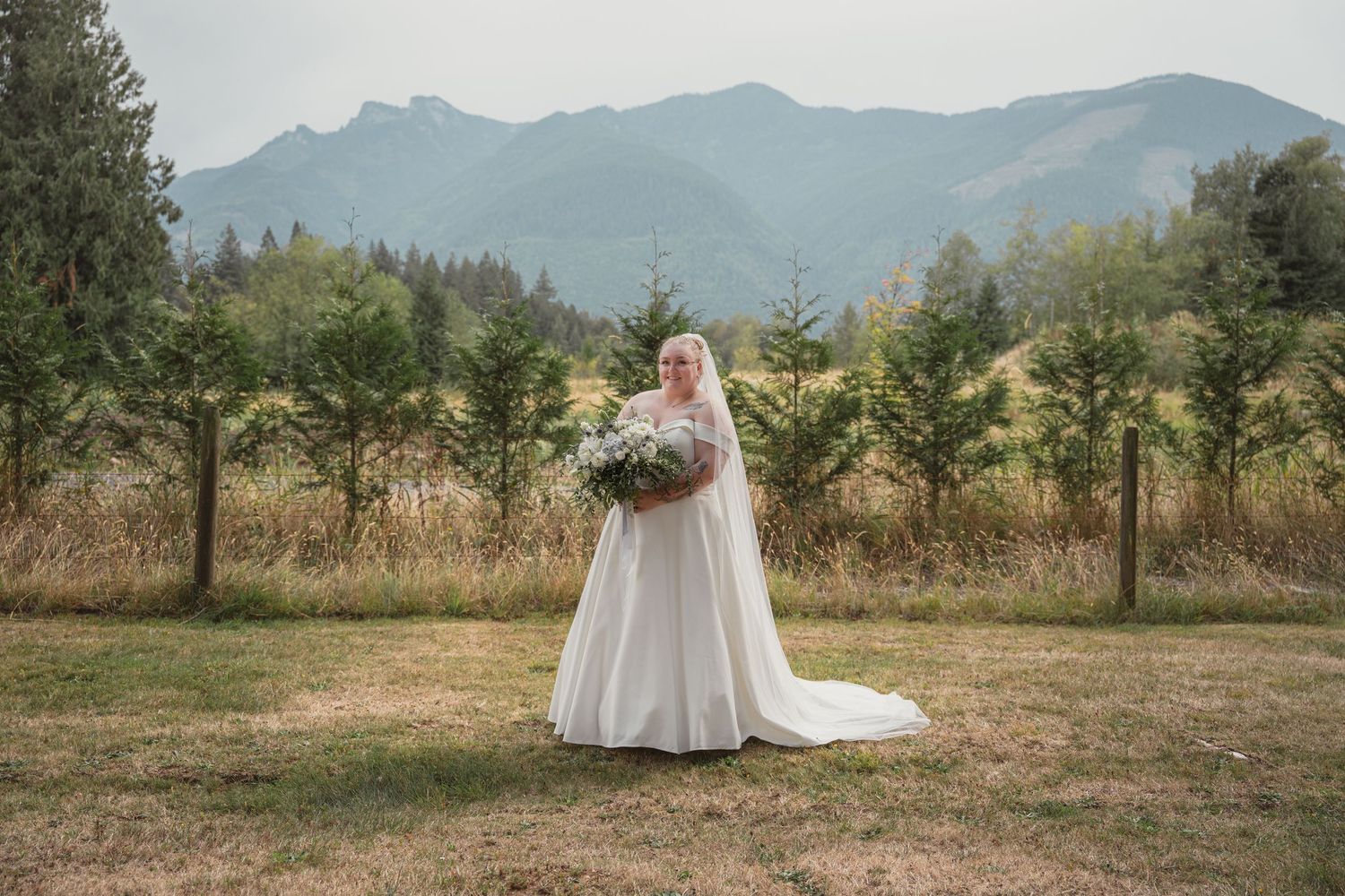 Bride in white gown and veil holds bouquet in mountain meadow with evergreen trees and misty peaks backdrop.