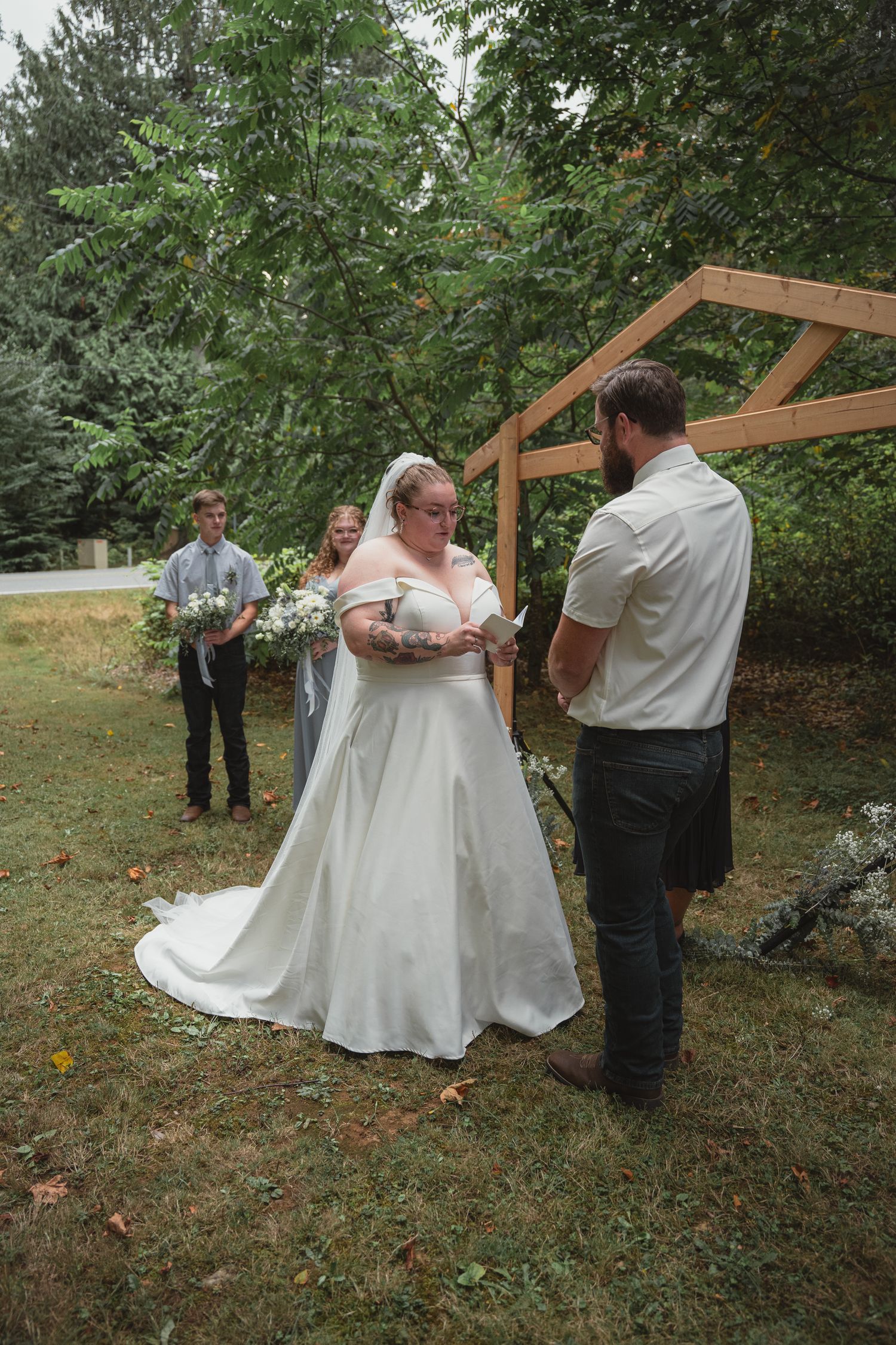 Bride reads vows to bearded groom at rustic wooden arch during intimate outdoor garden wedding ceremony.