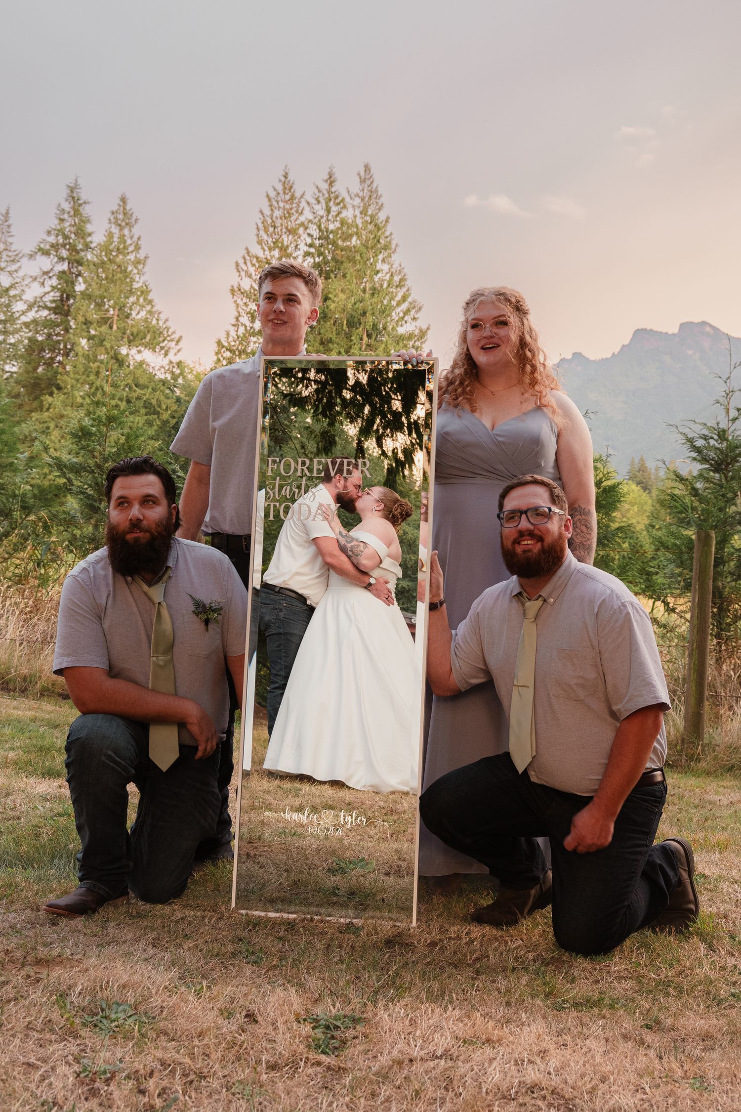 Wedding party of four holding a Forever sign with couple kissing photo outdoors near trees and mountains at sunset.