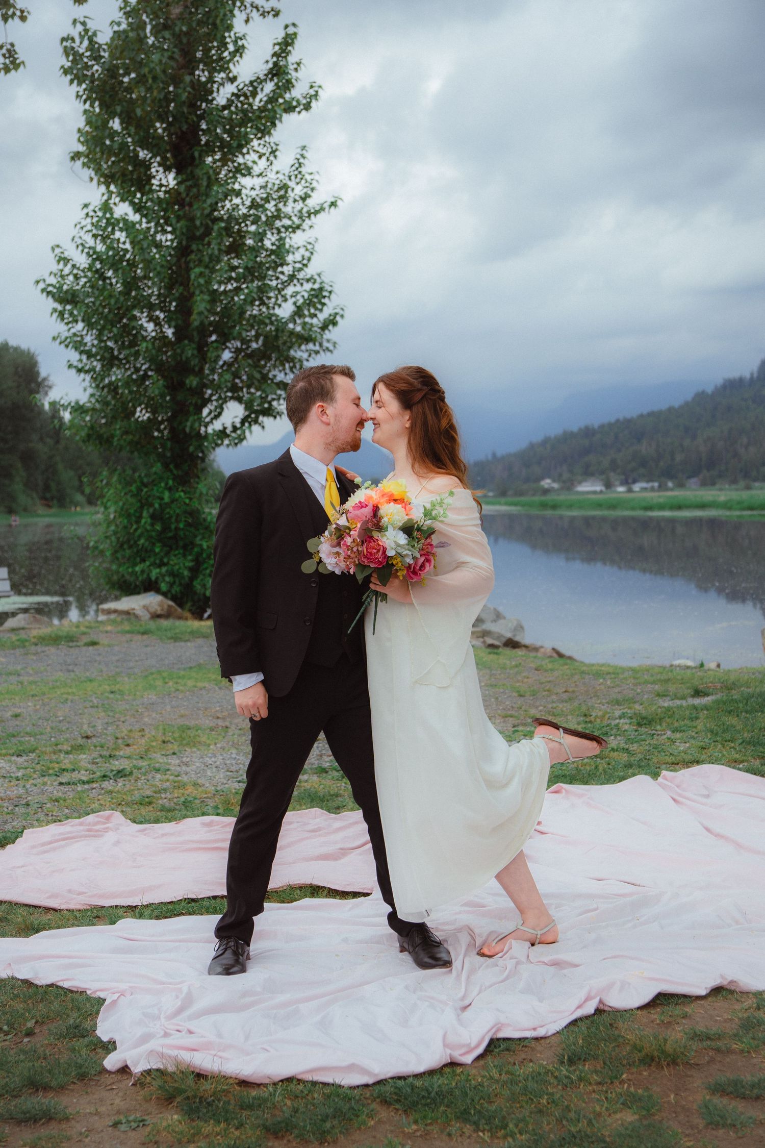 Bride and groom lean in for a kiss on pink fabric by a misty mountain lake.