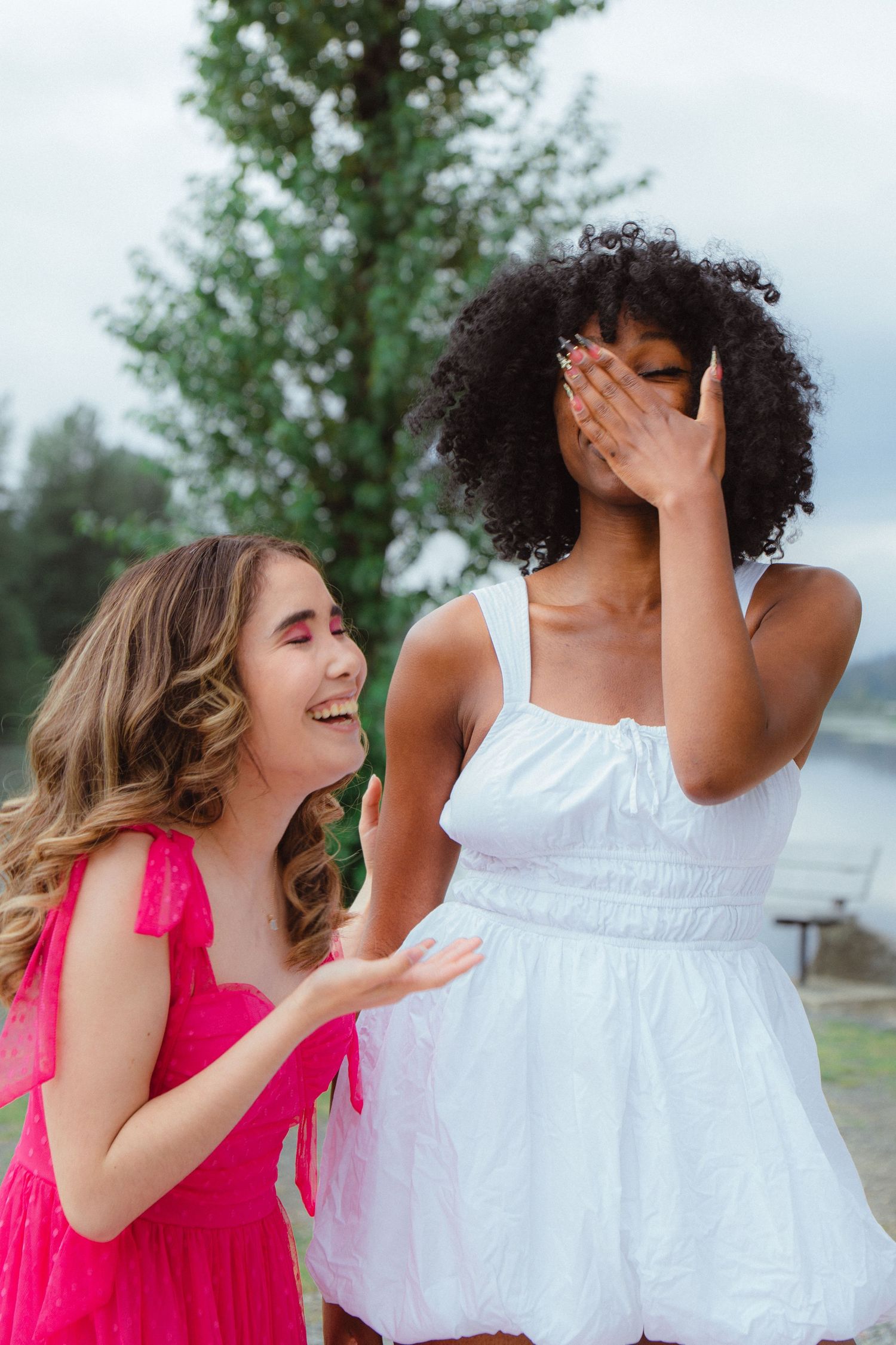 Two women in pink and white dresses laughing together outdoors near tall tree on overcast day.