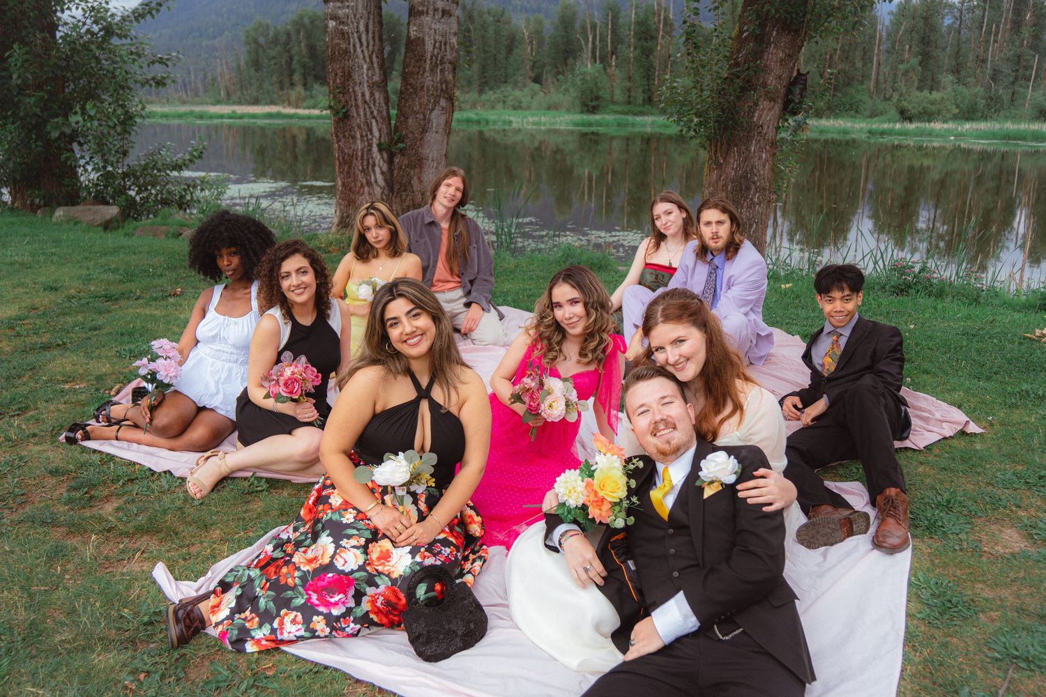 Happy wedding guests in colorful outfits lounging on blankets by a tranquil lakeside setting.