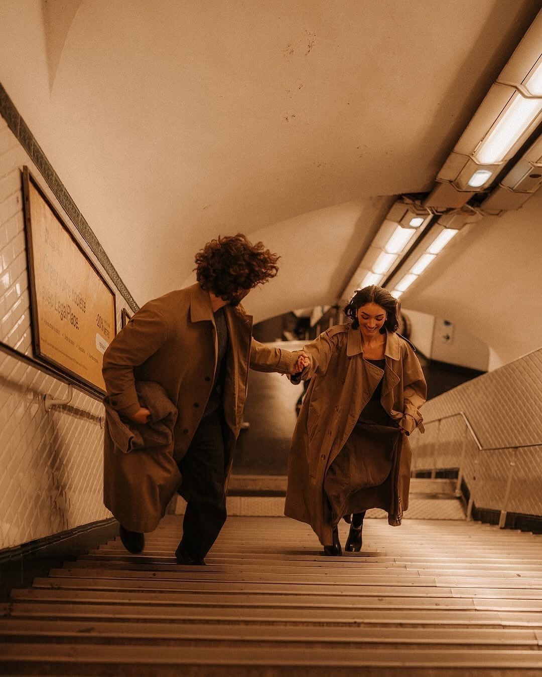 Couple in matching brown trench coats holding hands on subway station stairs.