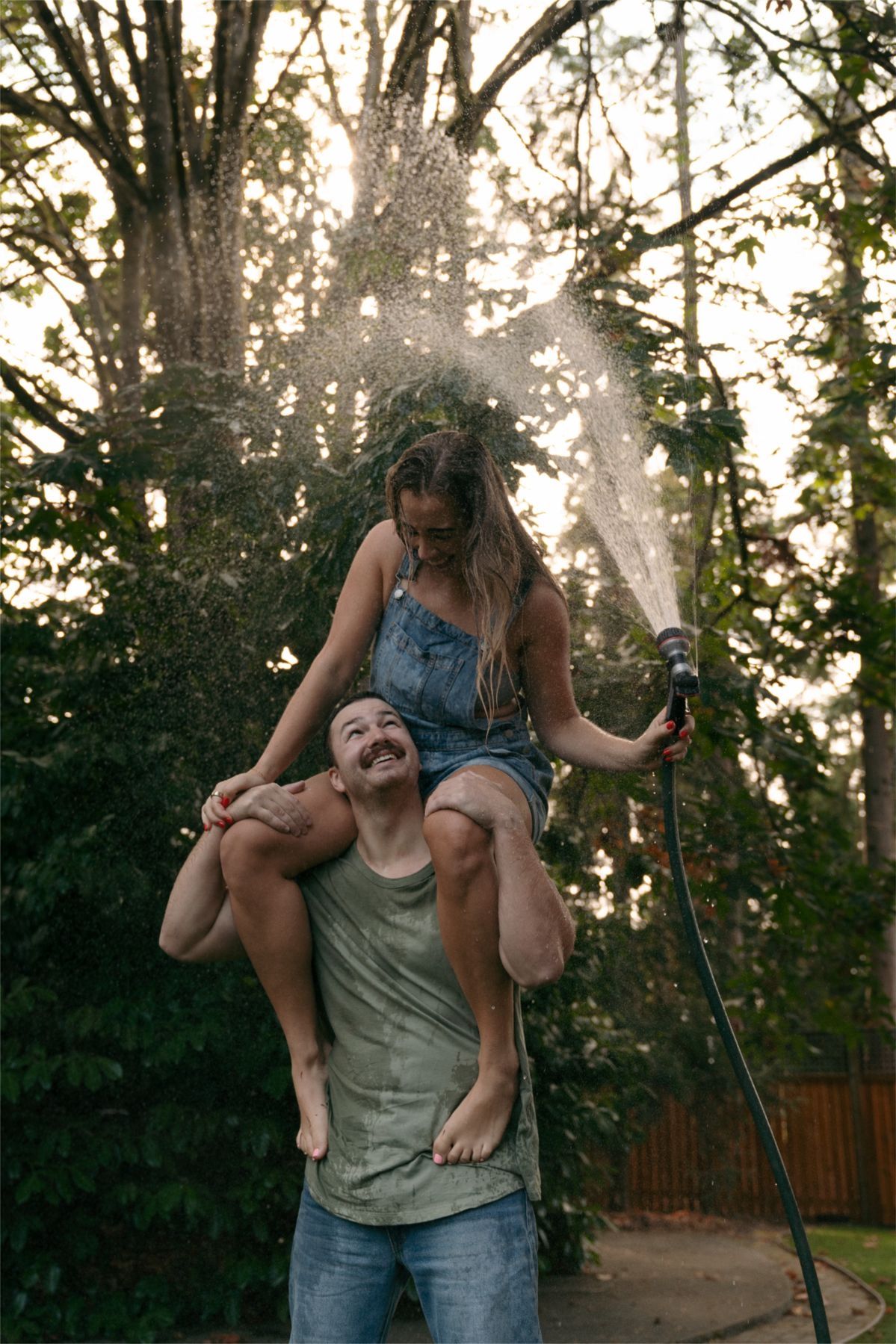 A laughing man carries a girl on his shoulders as she sprays them both with a garden hose outdoors.