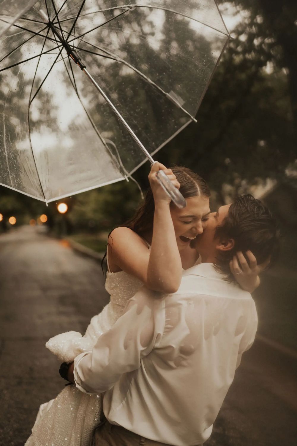 Couple sharing a romantic kiss in the rain under a clear umbrella, groom lifting bride in wedding attire outdoors.
