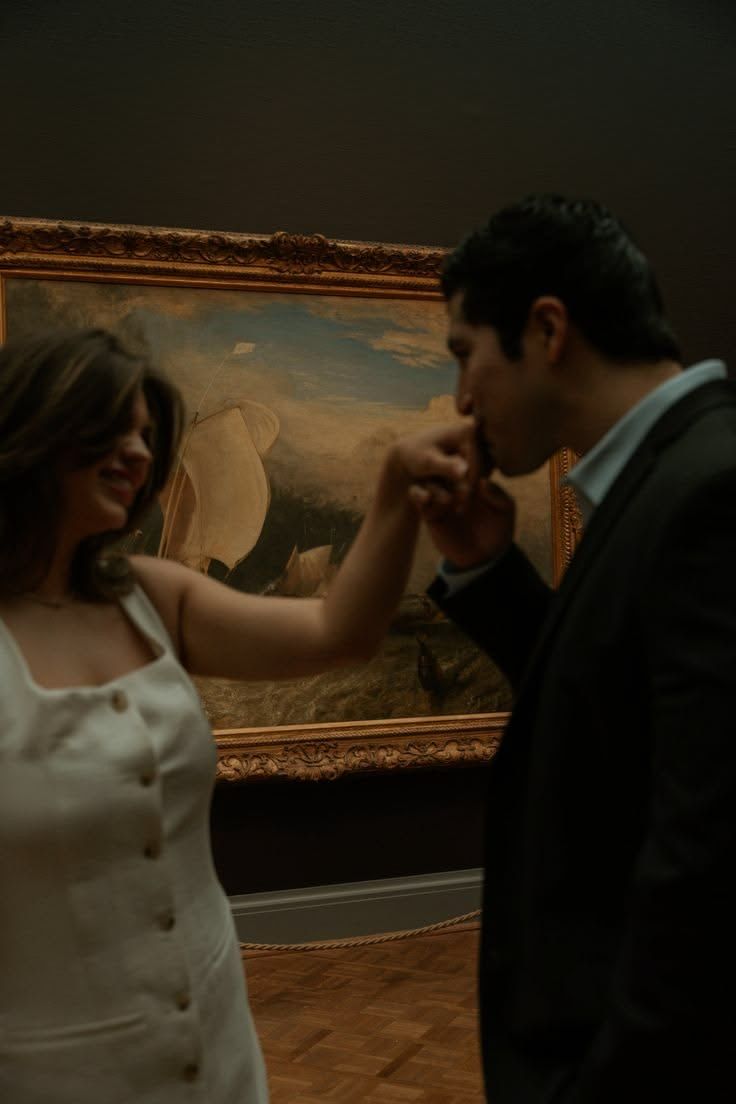 A man in a suit kisses a woman's hand in front of a framed painting in a dimly lit art museum.