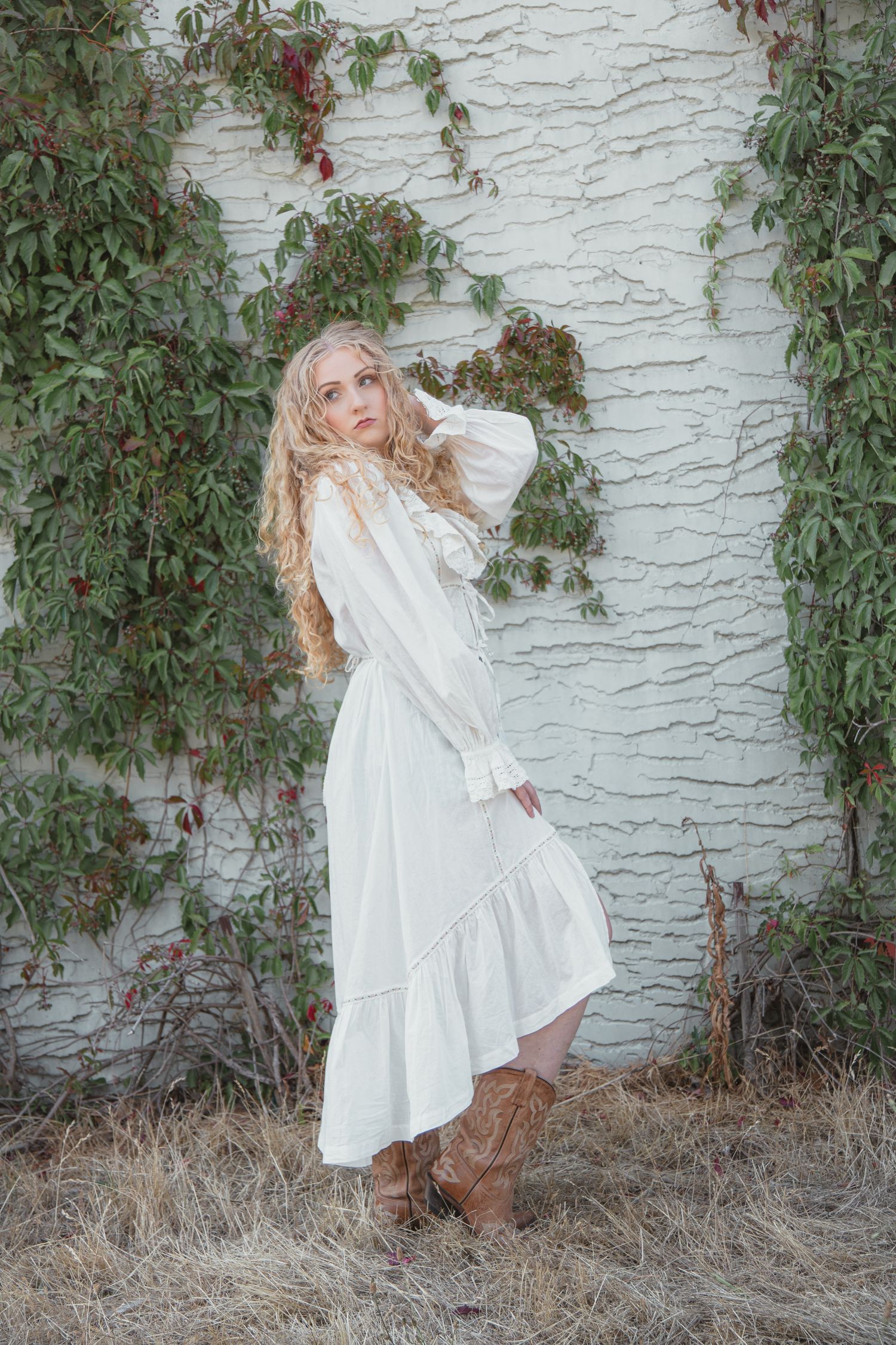 Blonde woman in white prairie dress and cowboy boots poses against a white brick wall with green ivy.