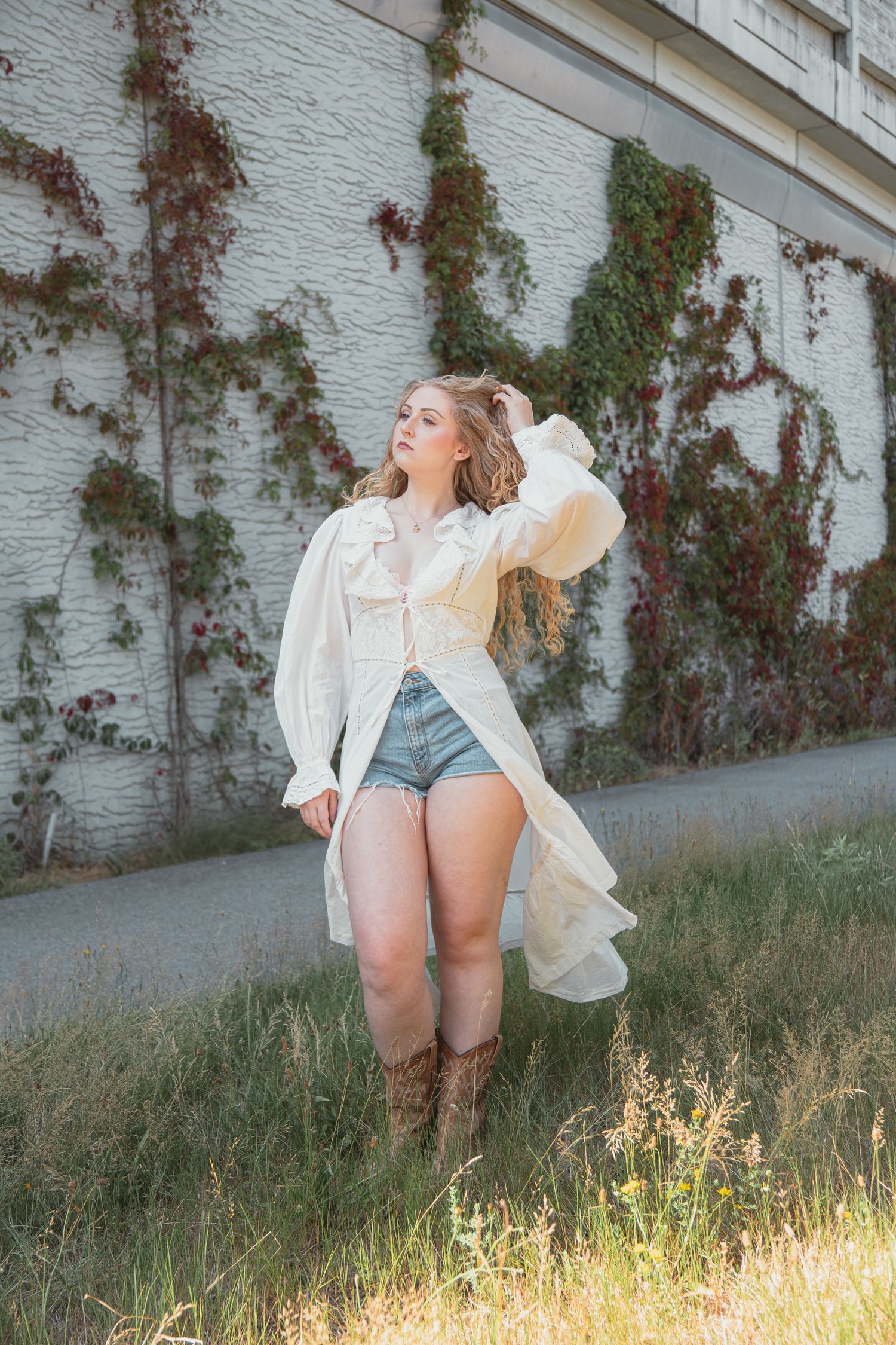 Blonde woman in white duster coat, denim shorts and cowboy boots posing by ivy-covered wall.