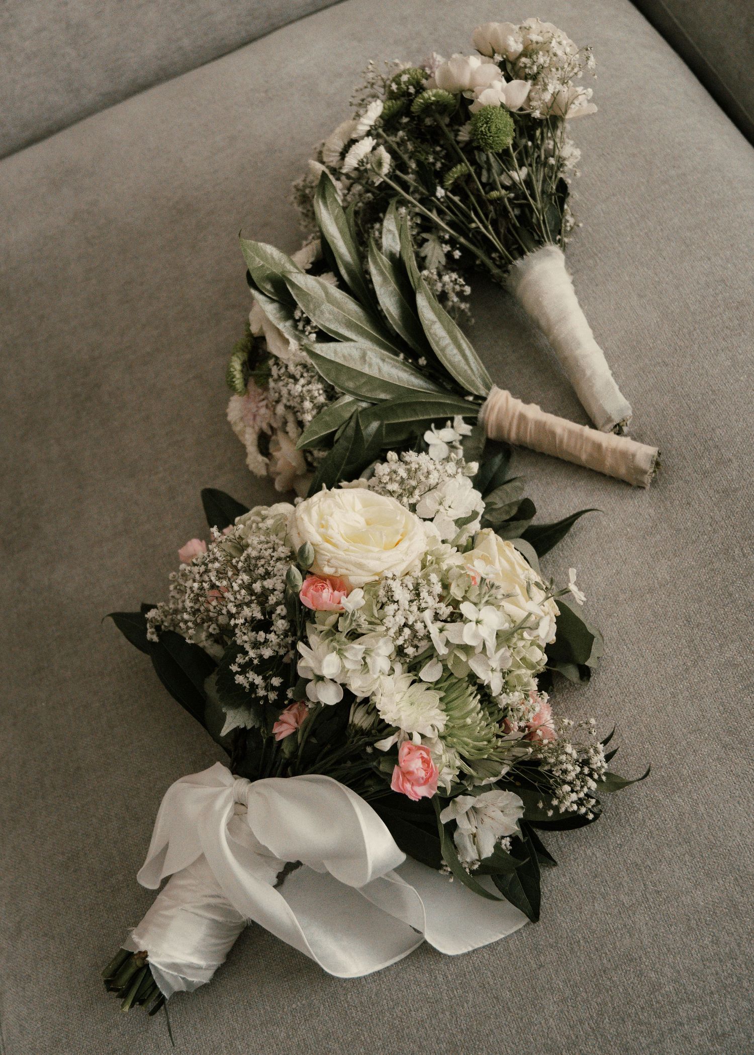 Three elegant wedding bouquets with white roses, baby's breath, and greenery arranged on a gray couch.