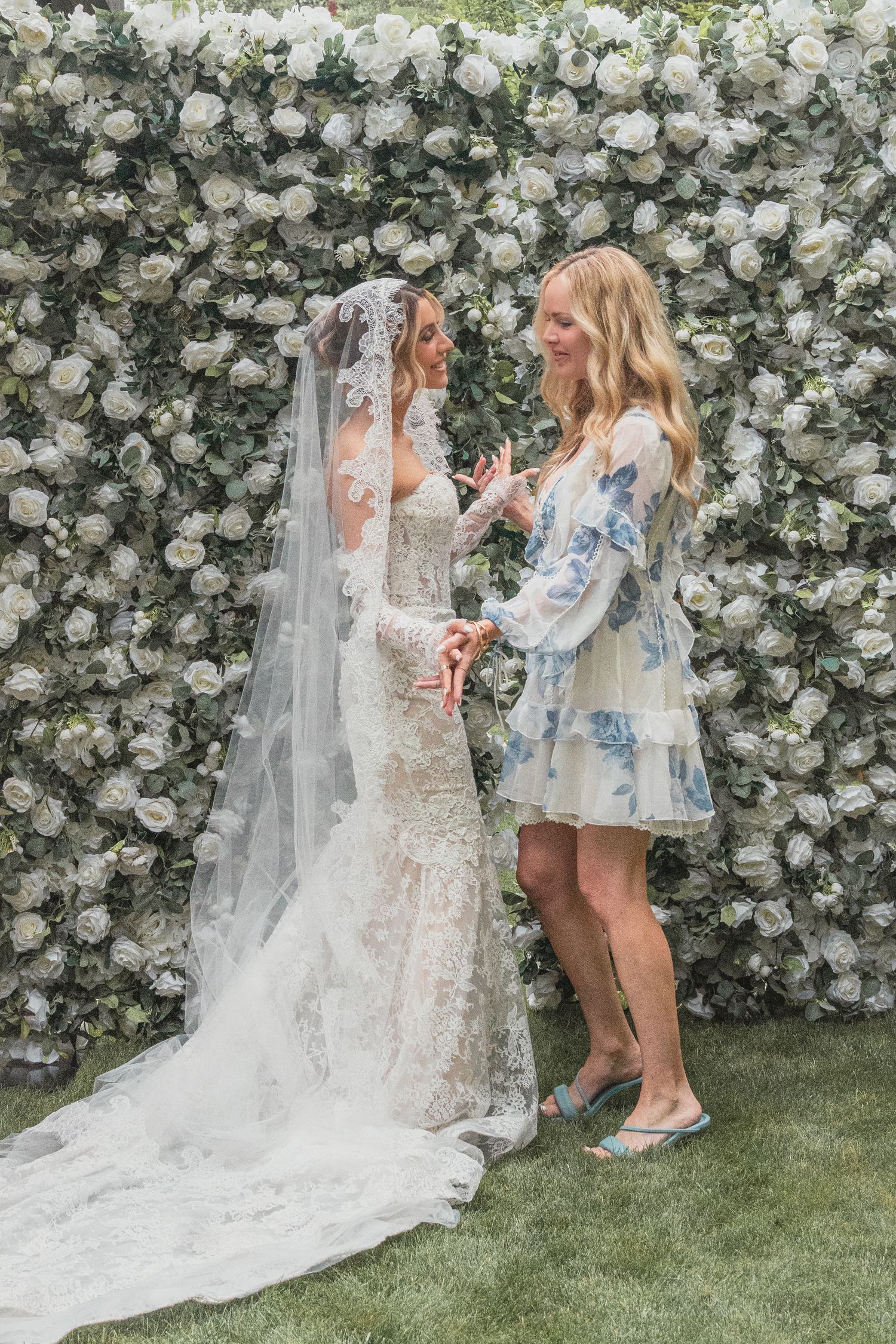 Bride in lace gown and veil with guest in floral dress, posing before white rose flower wall backdrop.