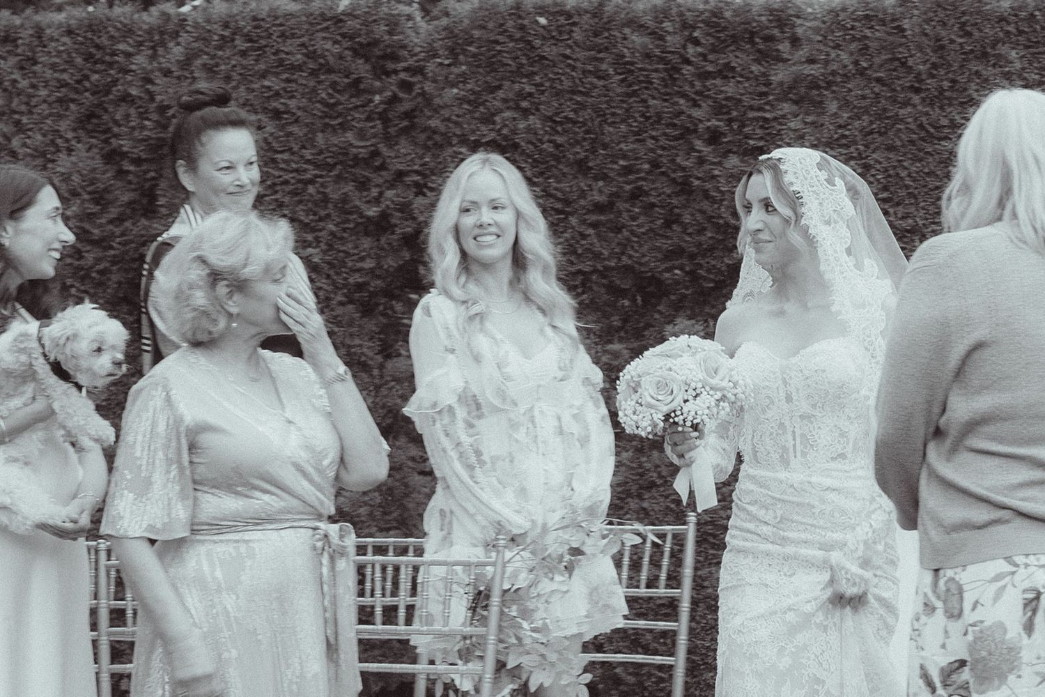 Black and white photo of a bride in veil holding bouquet with bridesmaids at outdoor wedding ceremony.