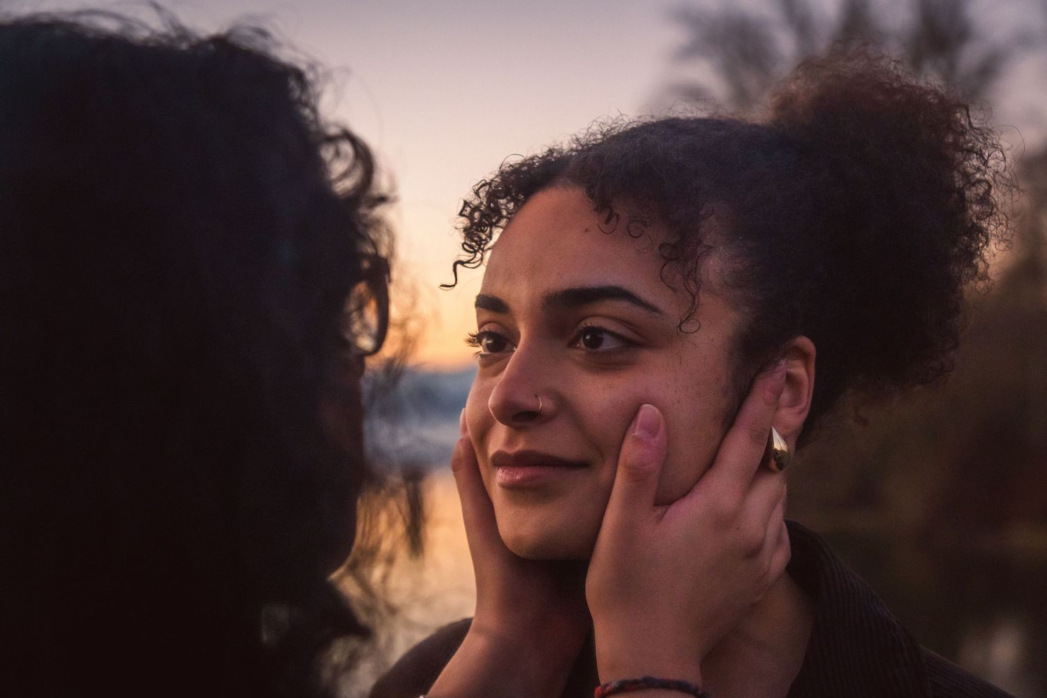 A woman smiles warmly as her partner gently cups her face during a beautiful golden hour sunset outdoors.