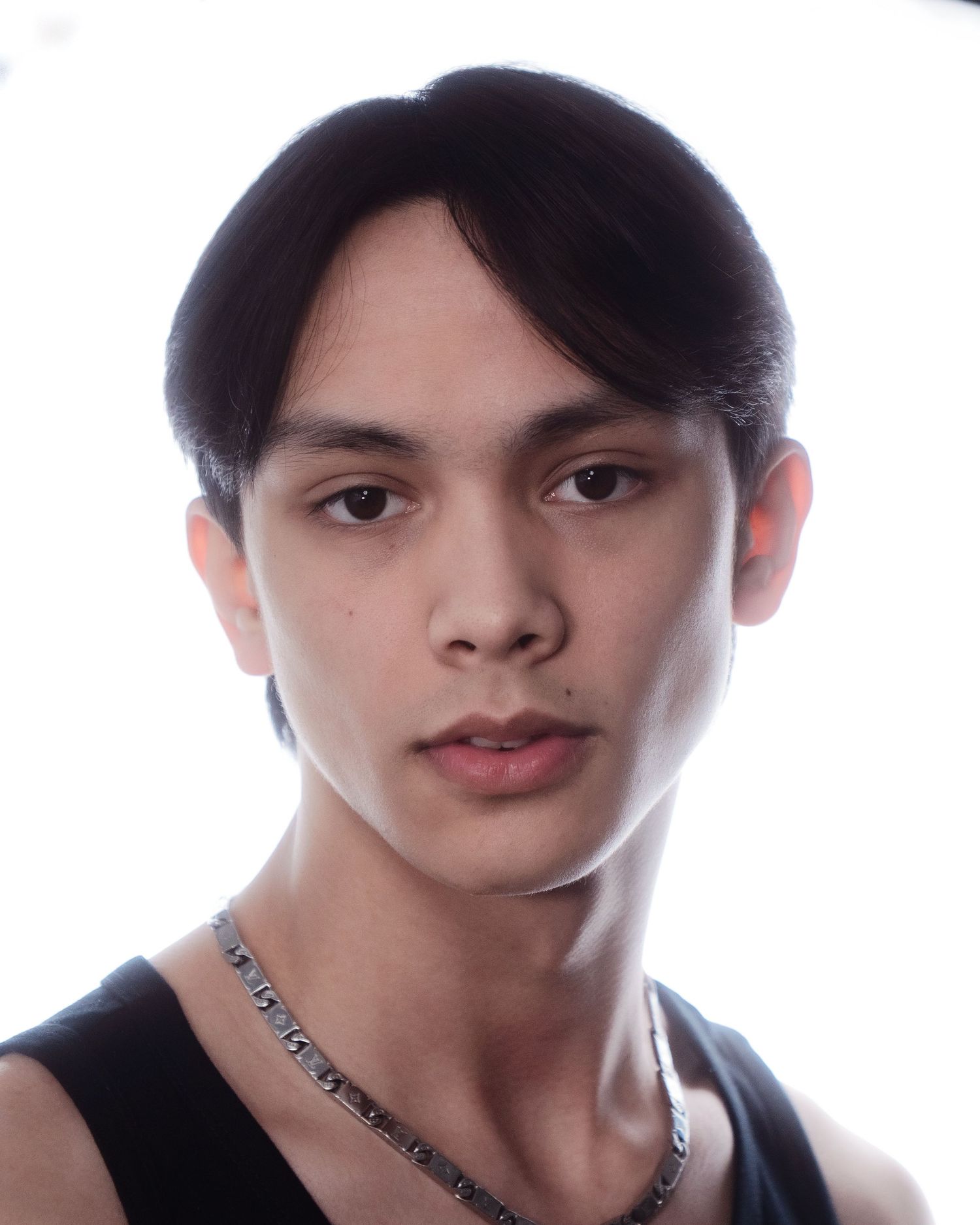 Young man with dark side-swept hair wearing a black tank top and chain necklace, portrait on white background.