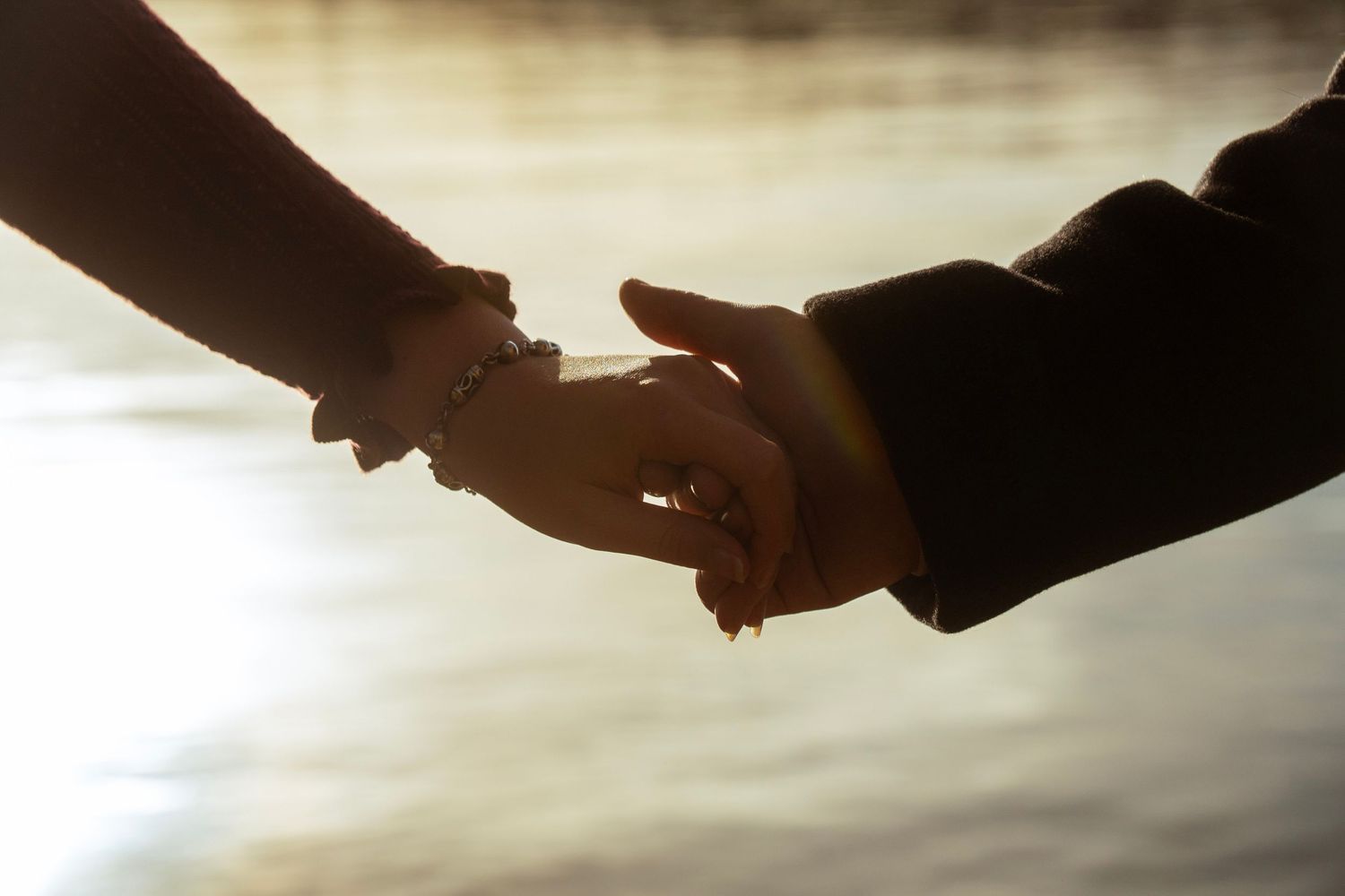 Romantic couple holding hands in silhouette against shimmering sunset water backdrop.