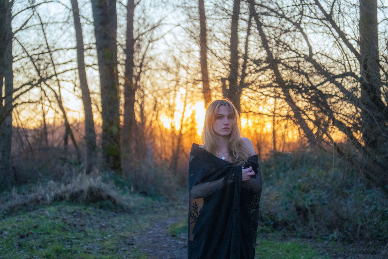 Blonde woman wrapped in a dark shawl standing in a forest with golden sunset behind her.