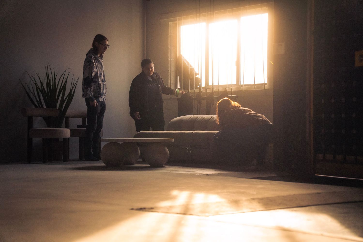 Three people arranging furniture on a sunlit film set with dramatic window light.