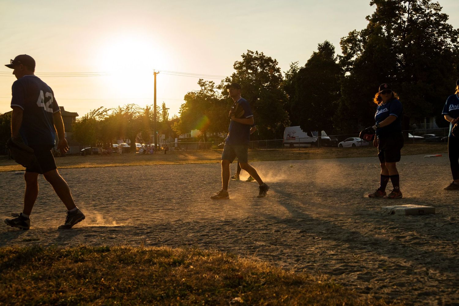 Players in blue uniforms run the bases on a dusty softball diamond during a golden hour game.