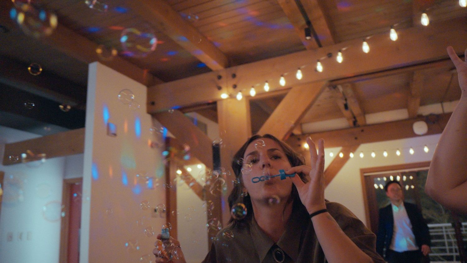 Guest blows bubbles at warmly lit wedding reception with string lights and wooden interior.