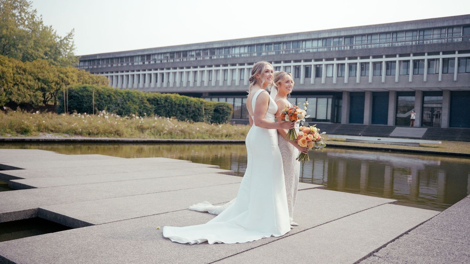 Two brides in white gowns hold bouquets outside a modern building with a reflecting pool.