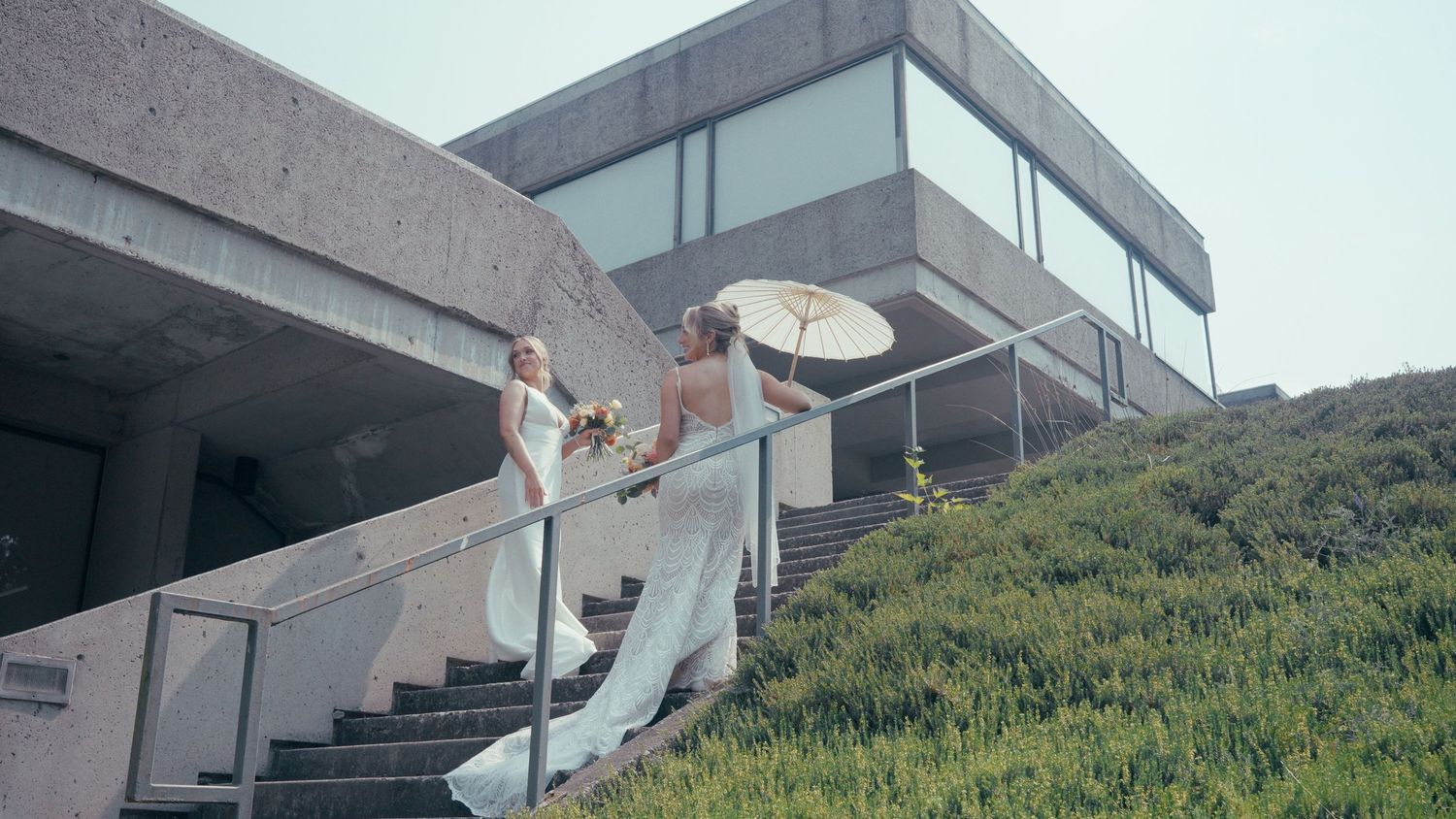 Two brides in flowing white gowns descend outdoor stairs near a modern concrete building with lush greenery.