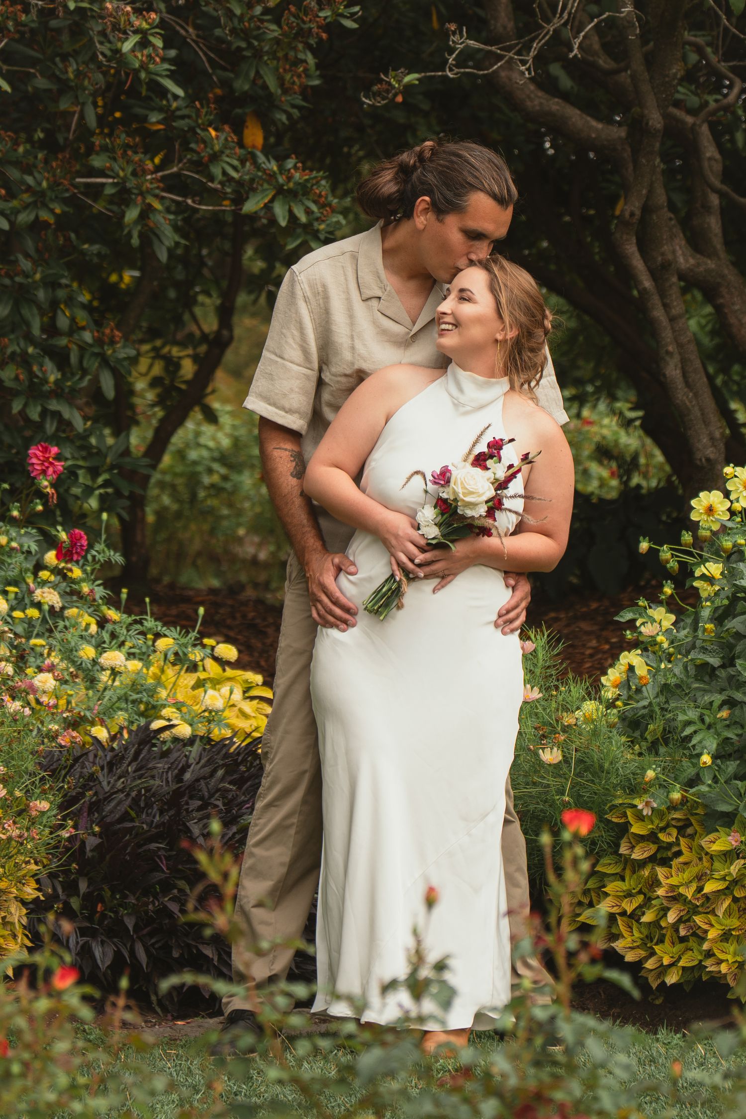 Bride in white halter gown holds bouquet as groom in tan suit kisses her forehead in a lush garden.