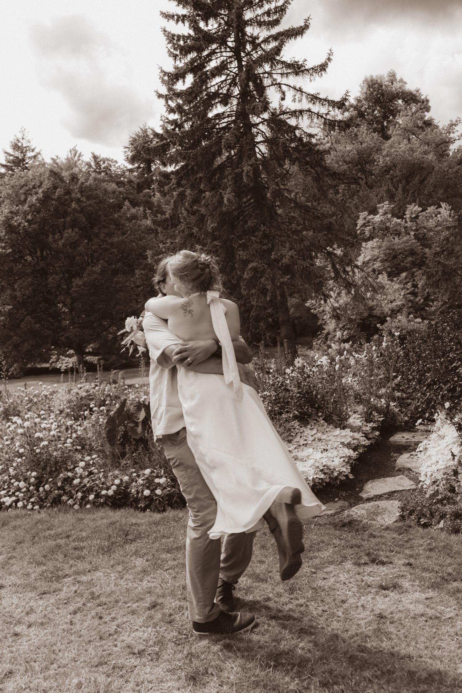 Sepia-toned photo of groom lifting bride in a beautiful garden setting with tall evergreen trees.