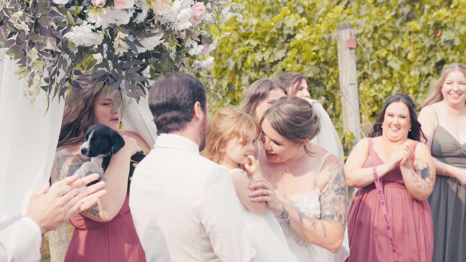 Bride and groom share an emotional embrace with wedding guests and bridesmaids in mauve dresses at an outdoor ceremony.