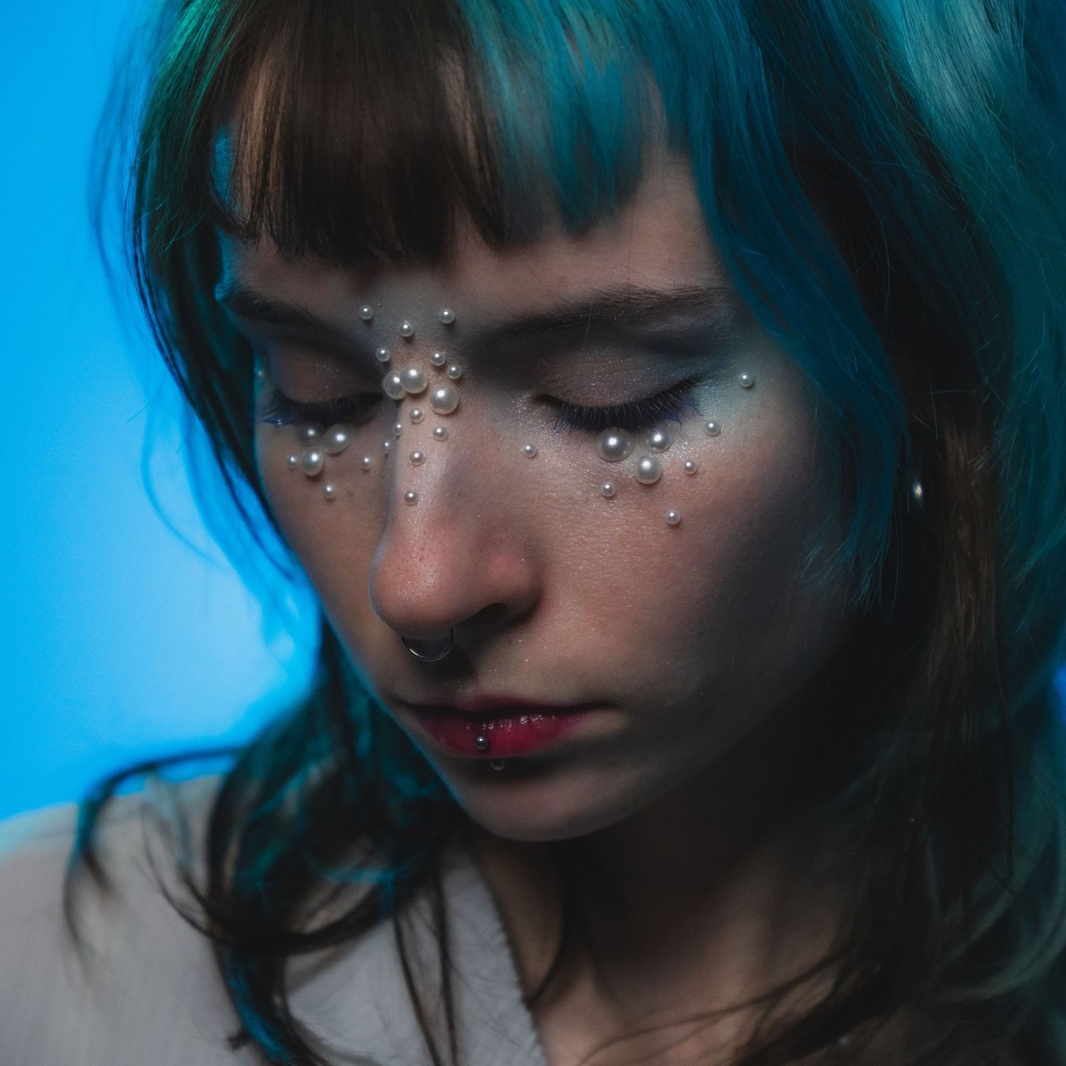 Woman with teal hair and pearl face gems looking down against a blue background.