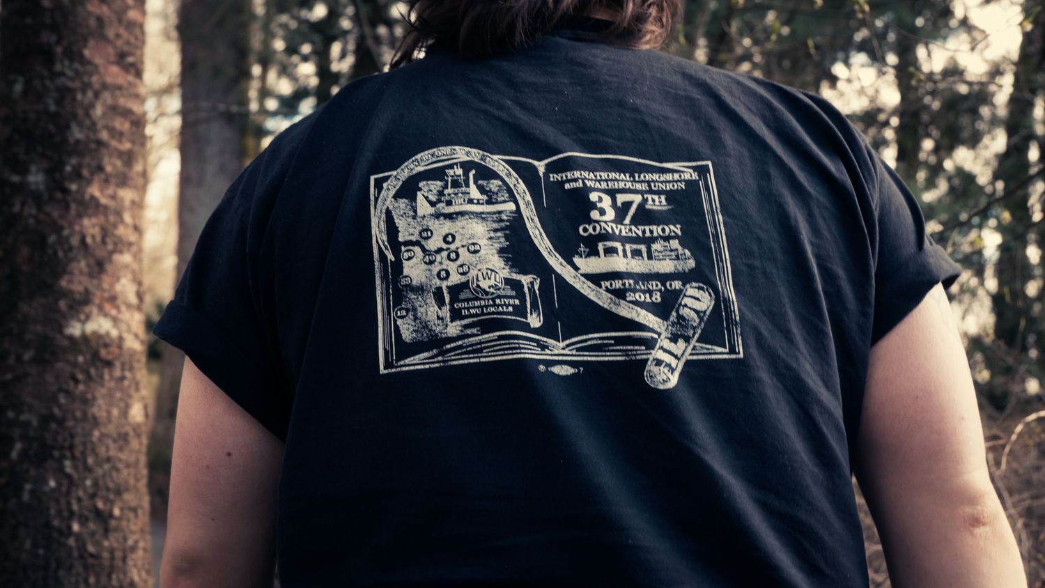 Back view of a person wearing a black t-shirt with a 37th convention graphic print in a forest.