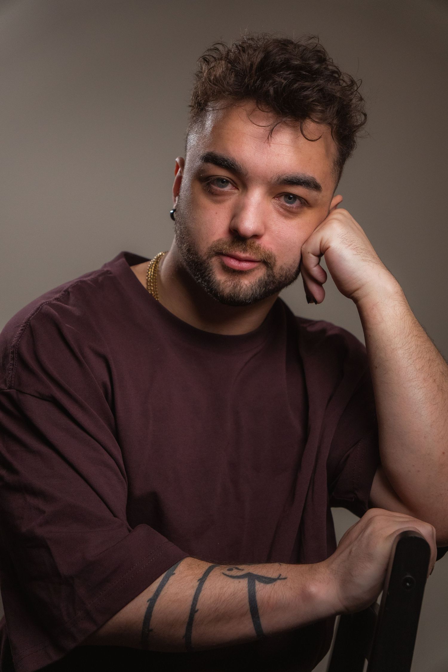 Bearded man in burgundy shirt with arm tattoo resting head on hand against gray background.