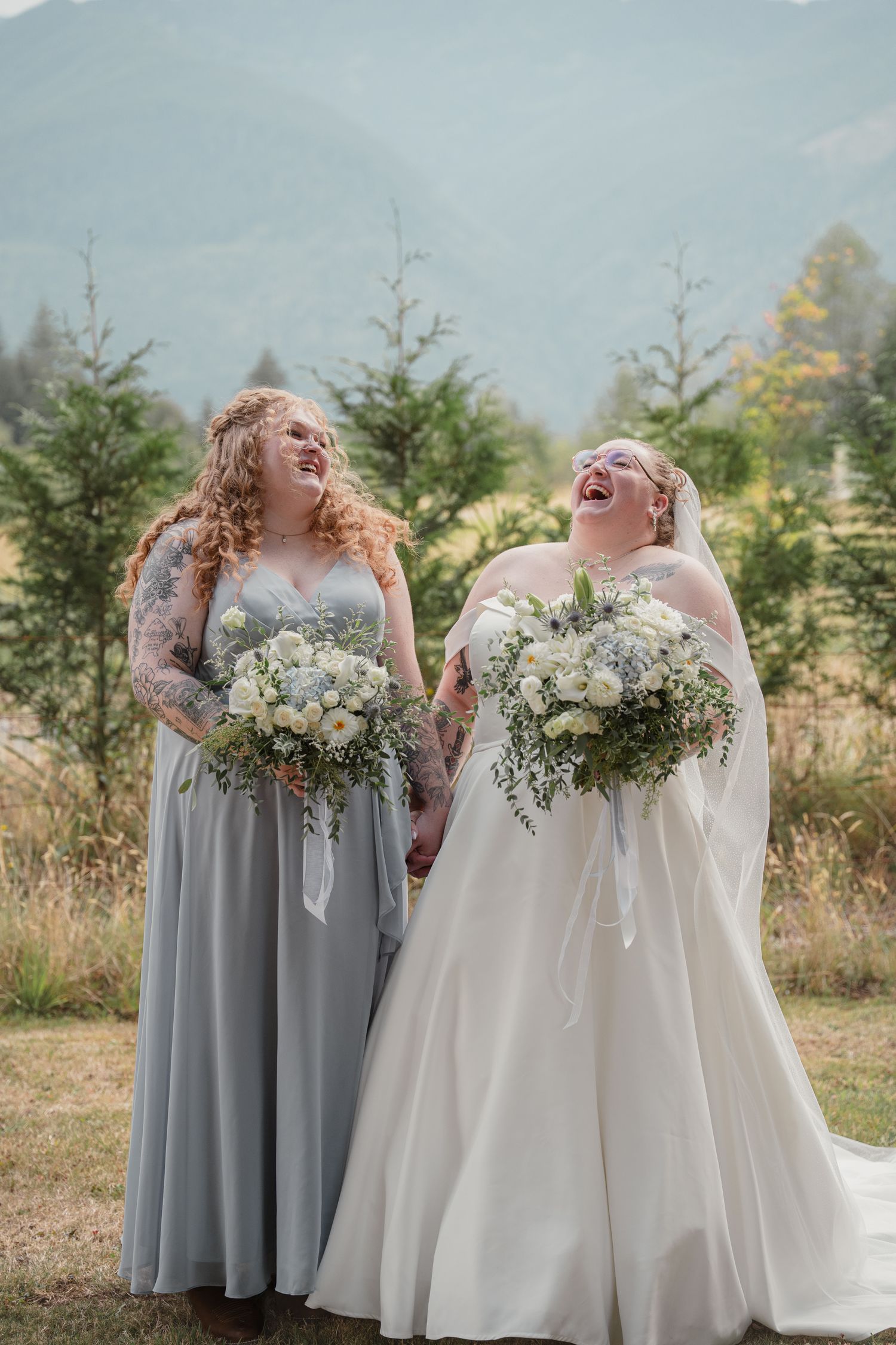 Bride and bridesmaid in gray dress laughing while holding bouquets with mountain backdrop.