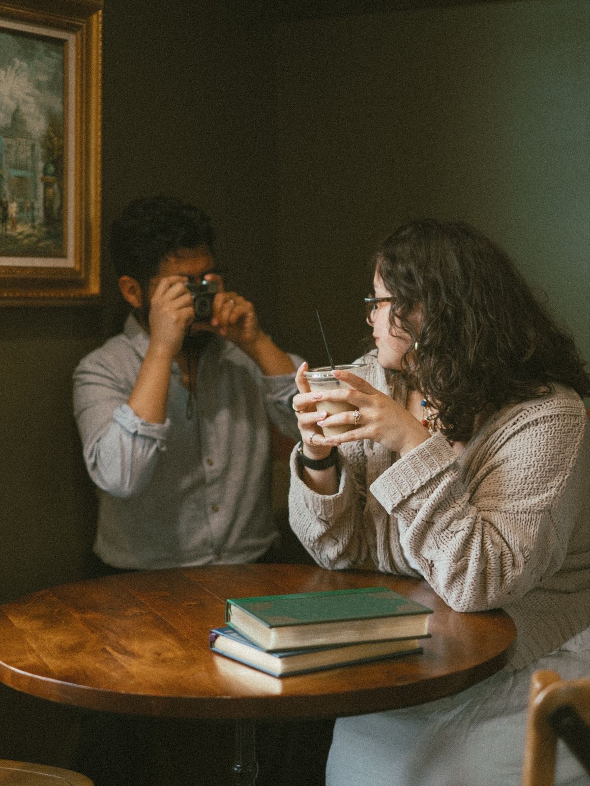 A woman sips coffee at a cafe table while a man photographs her beside stacked books and framed wall art.