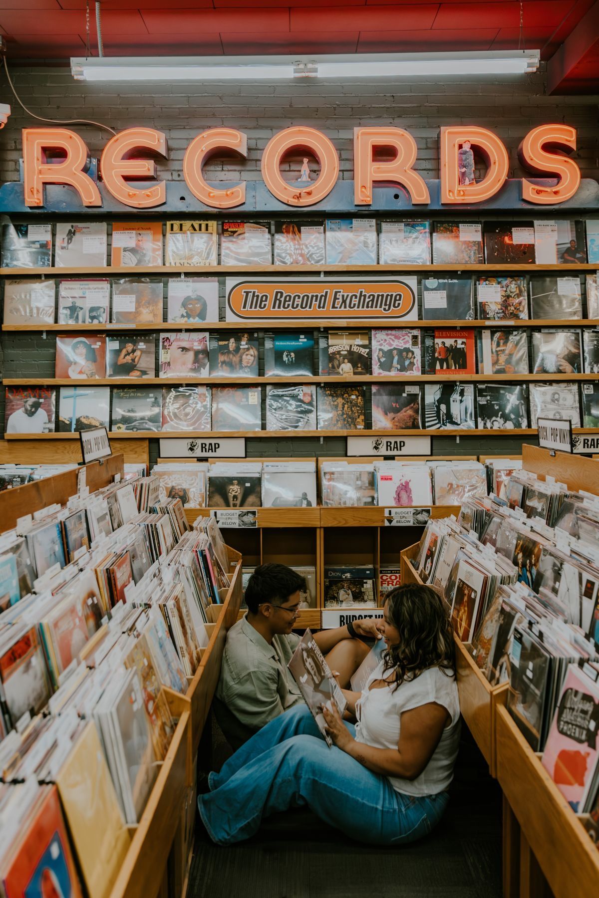 Couple sitting on floor browsing vinyl records at The Record Exchange store with illuminated RECORDS sign above.