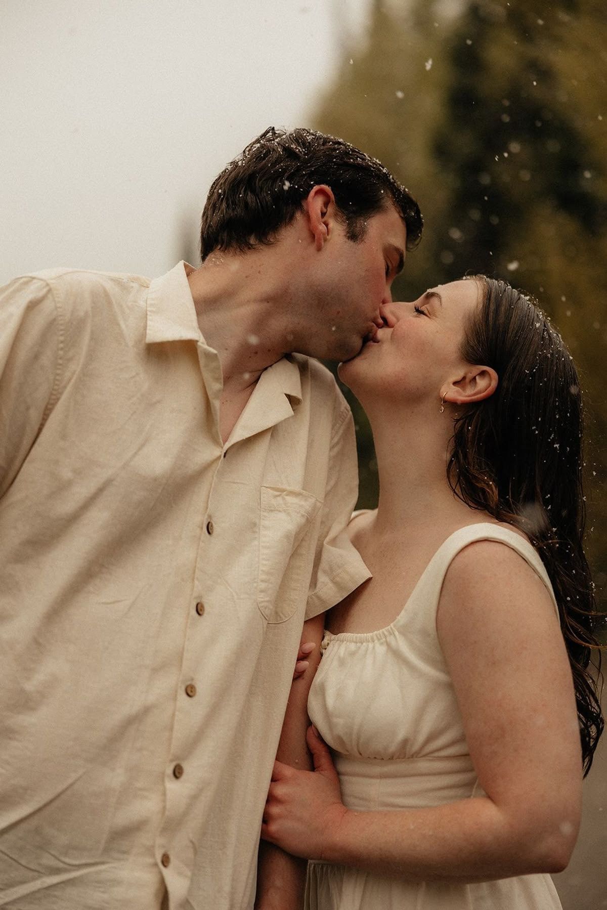 A couple shares a romantic kiss in the rain, dressed in matching cream outfits, embracing tenderly outdoors.