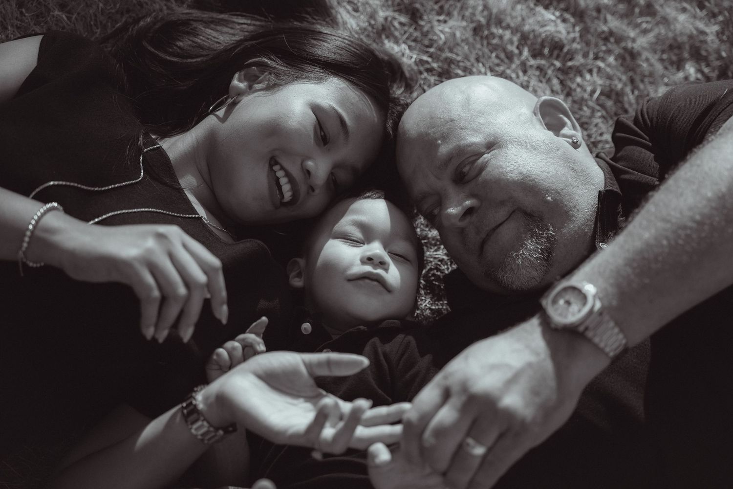Black and white photo of smiling parents lying on grass with their sleeping newborn baby between them.