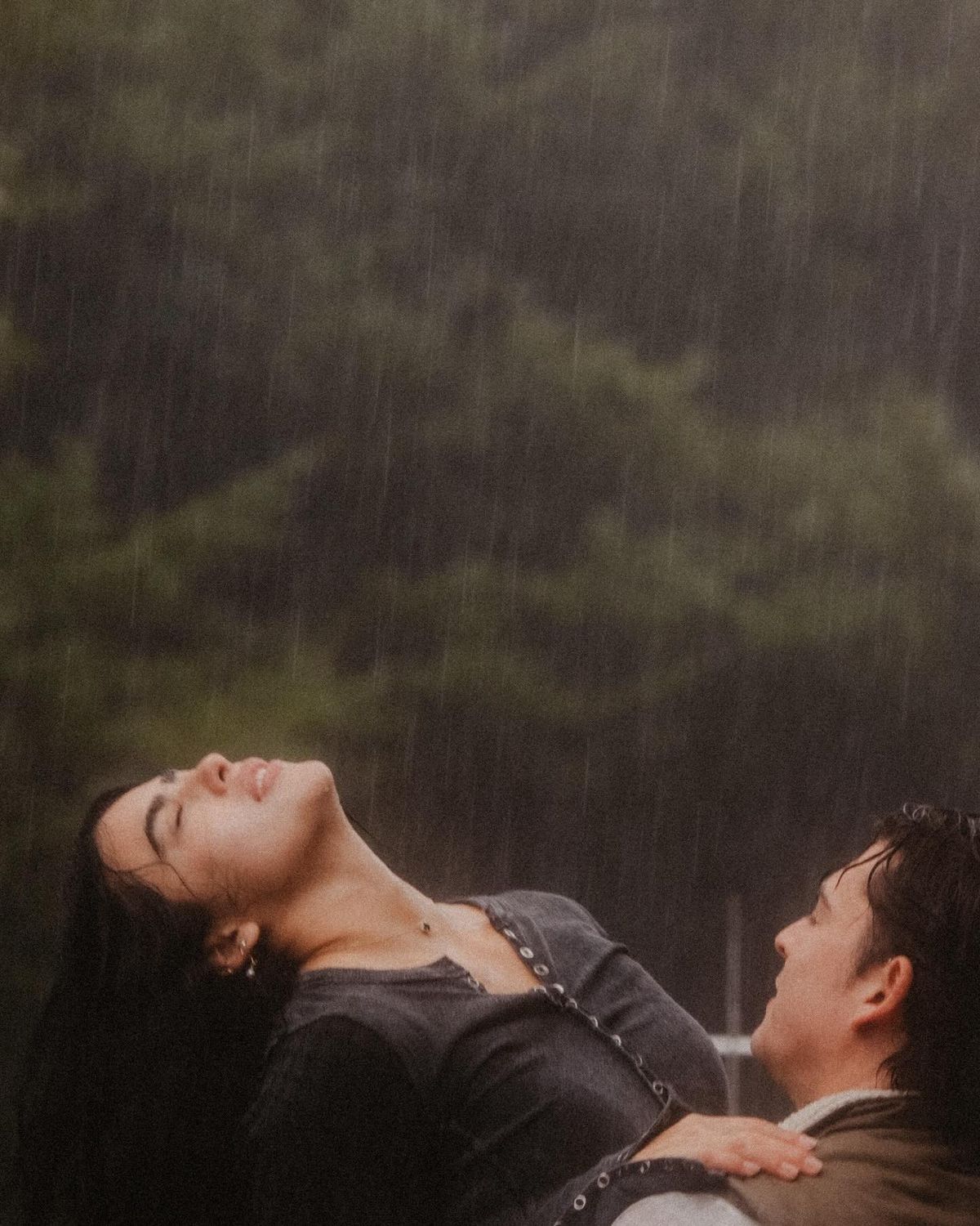 Two people share an emotional moment in heavy rain surrounded by lush green trees.