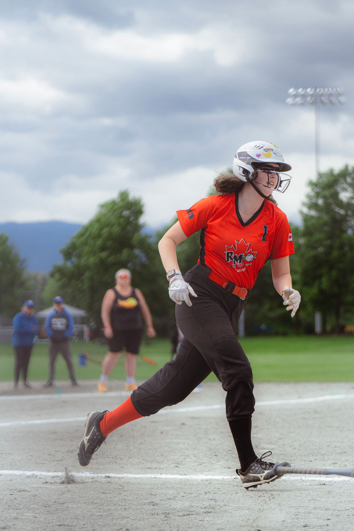 Female softball player in orange RMO jersey runs the bases smiling during an outdoor game.