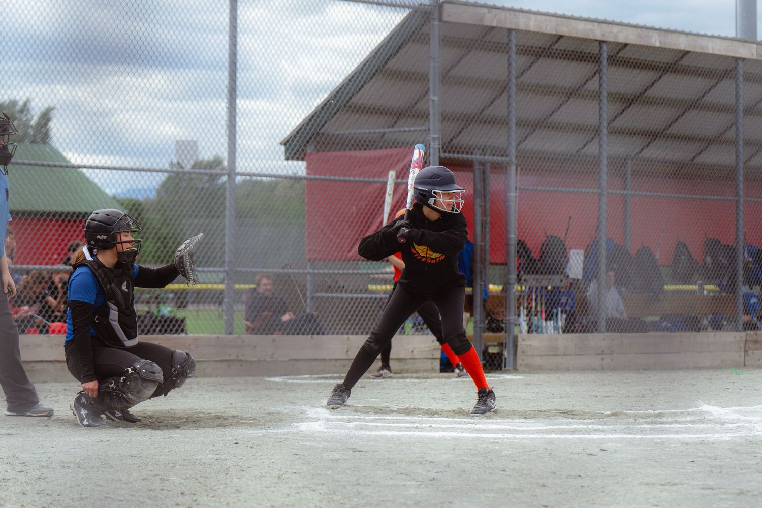Female softball batter in black uniform at home plate with catcher behind her.