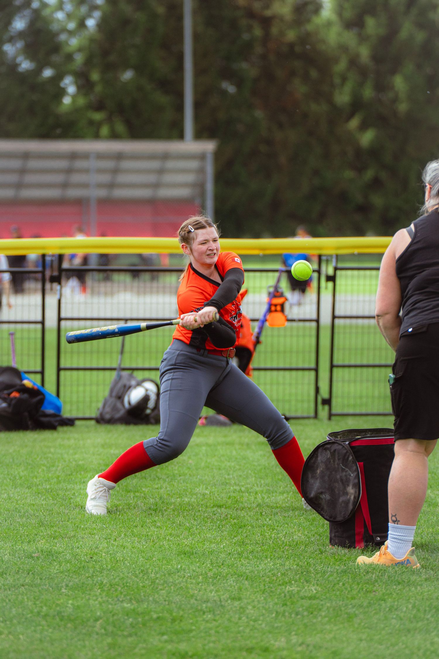 Female softball player in orange uniform swinging at yellow ball during outdoor batting practice.