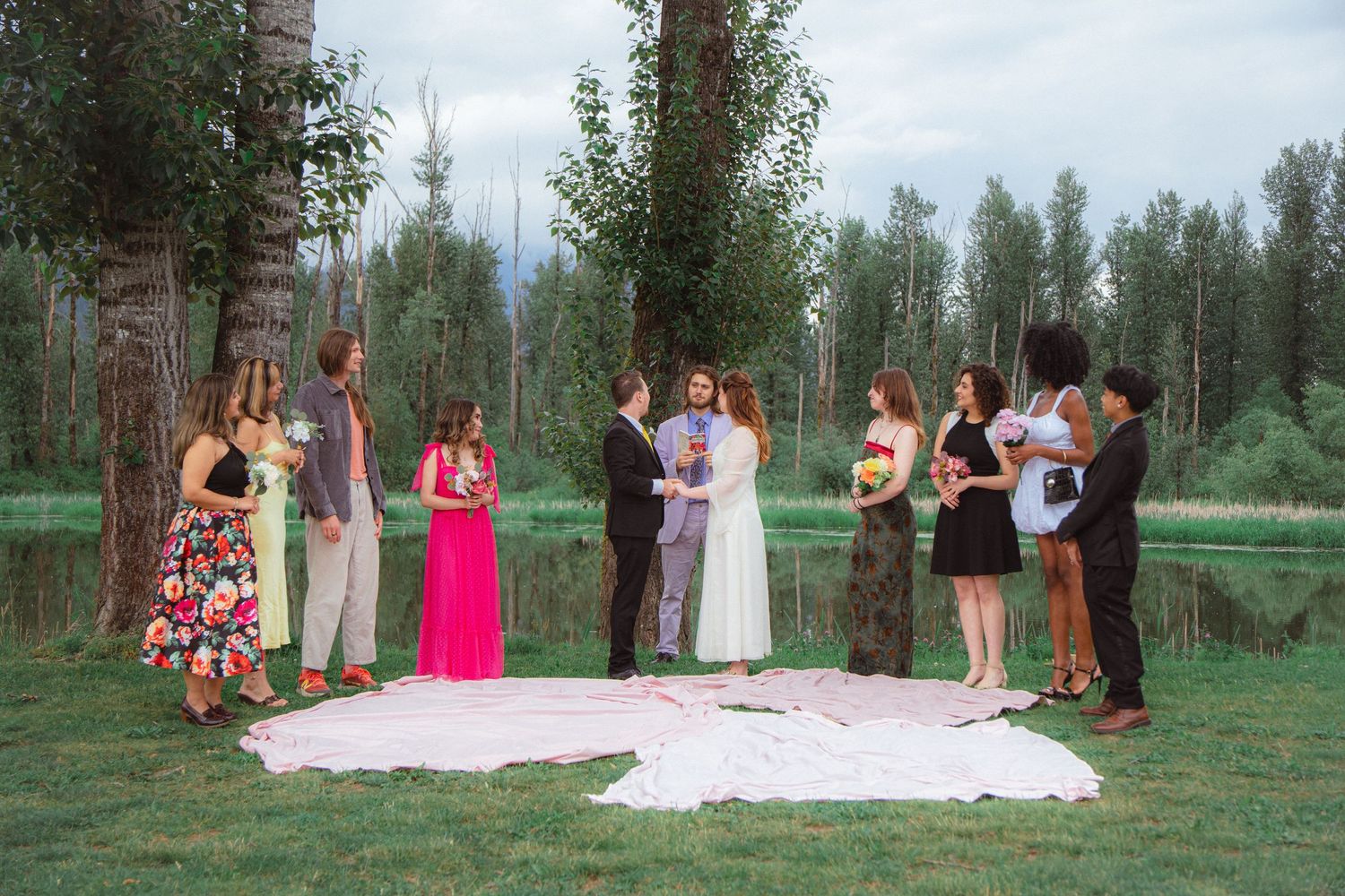 Small intimate wedding ceremony with guests gathered on a white blanket beside a tree-lined lake.