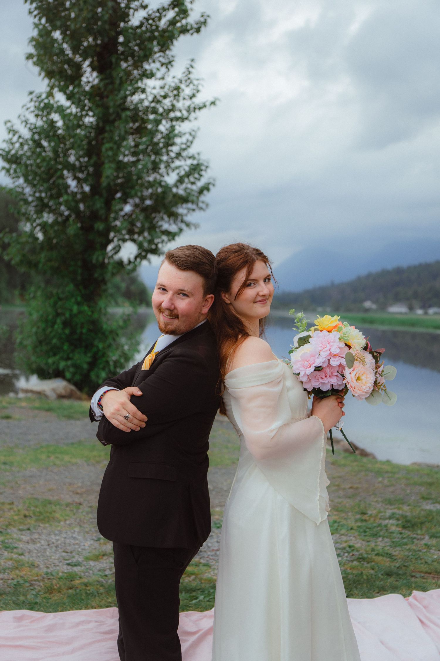 Bride and groom stand back-to-back smiling by a tree-lined mountain lake.