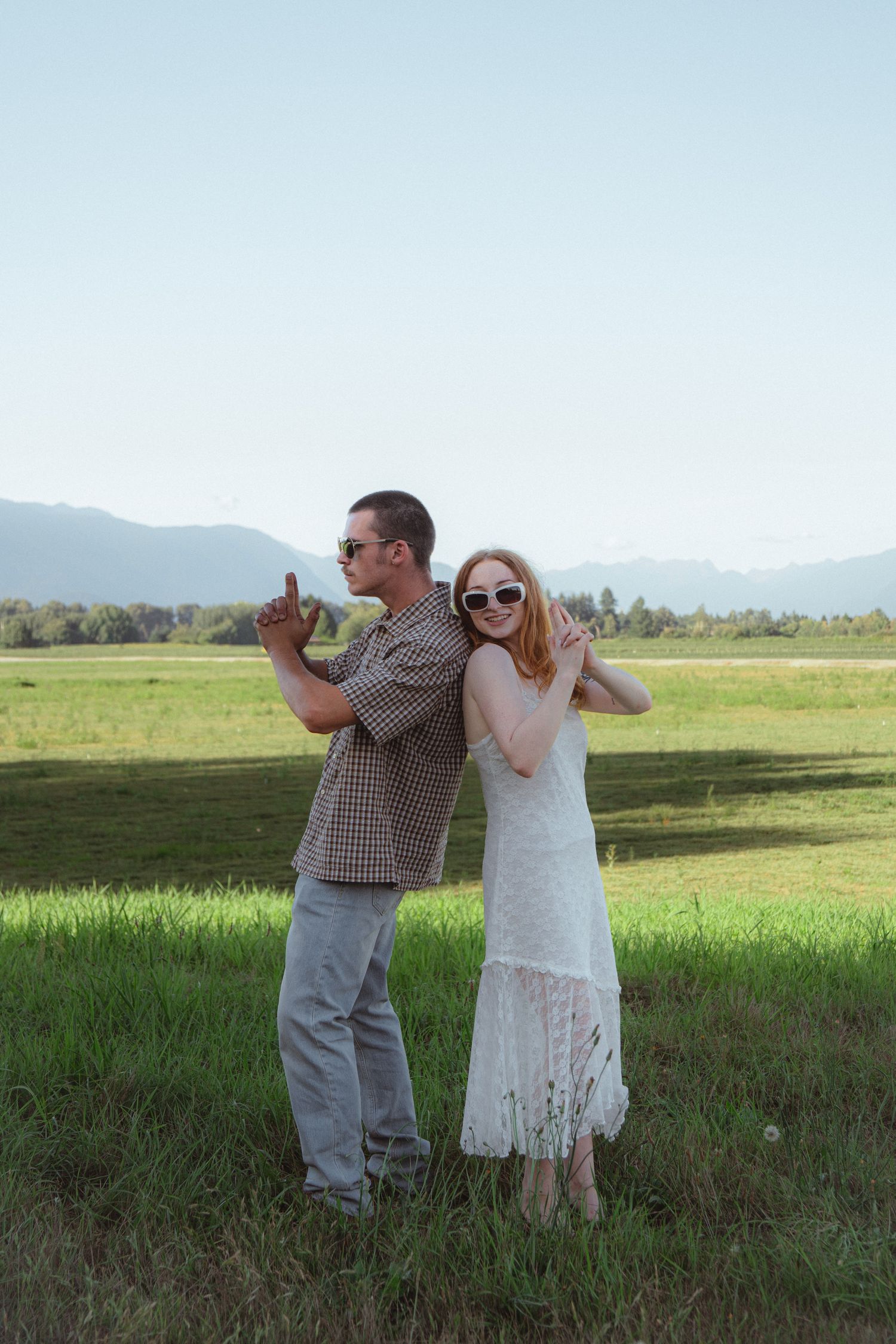 A couple in sunglasses posing in an open green field with mountains in the background on a sunny day.