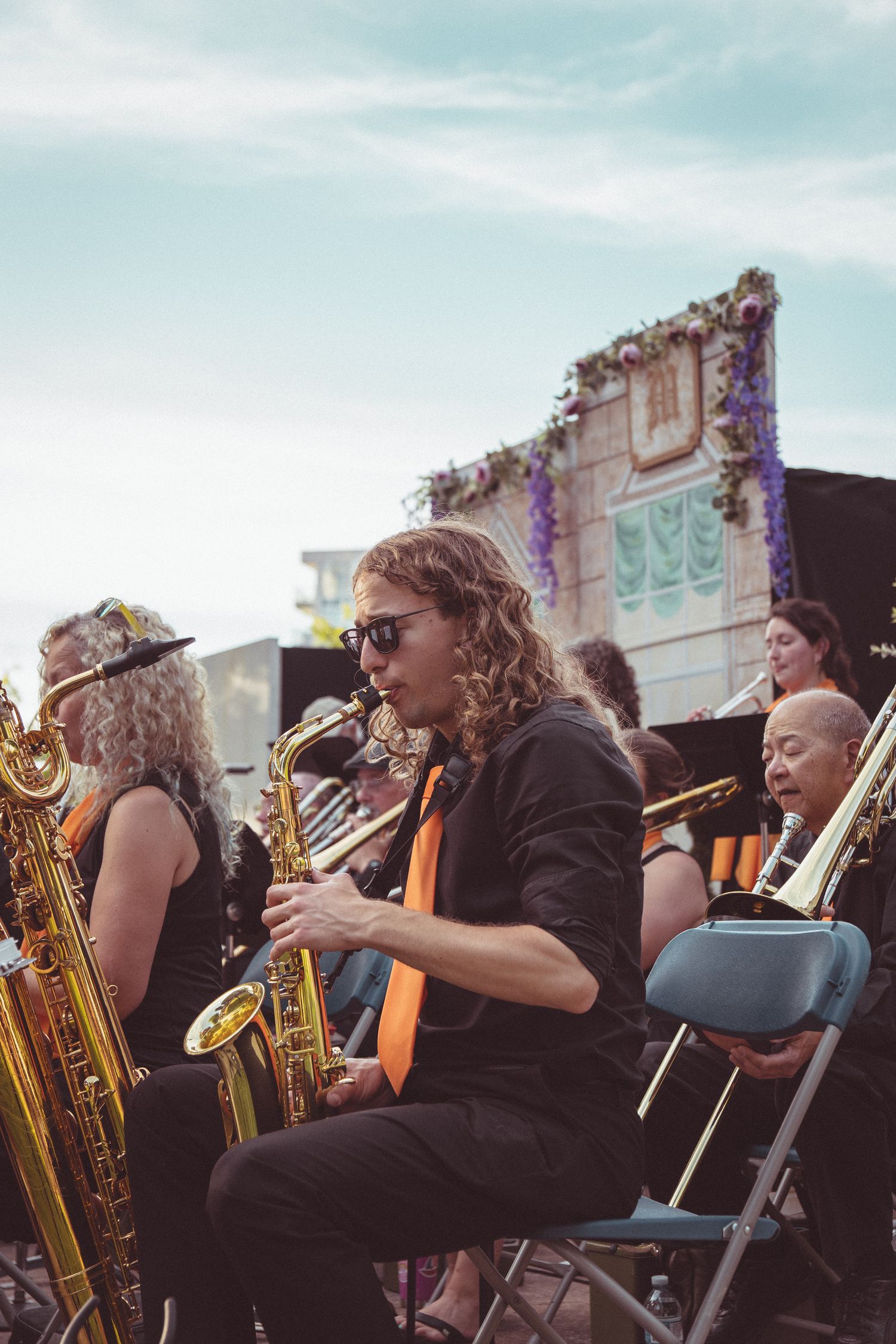 Musicians playing saxophones and brass instruments outdoors at a festival, led by a curly-haired man in sunglasses and orange tie.