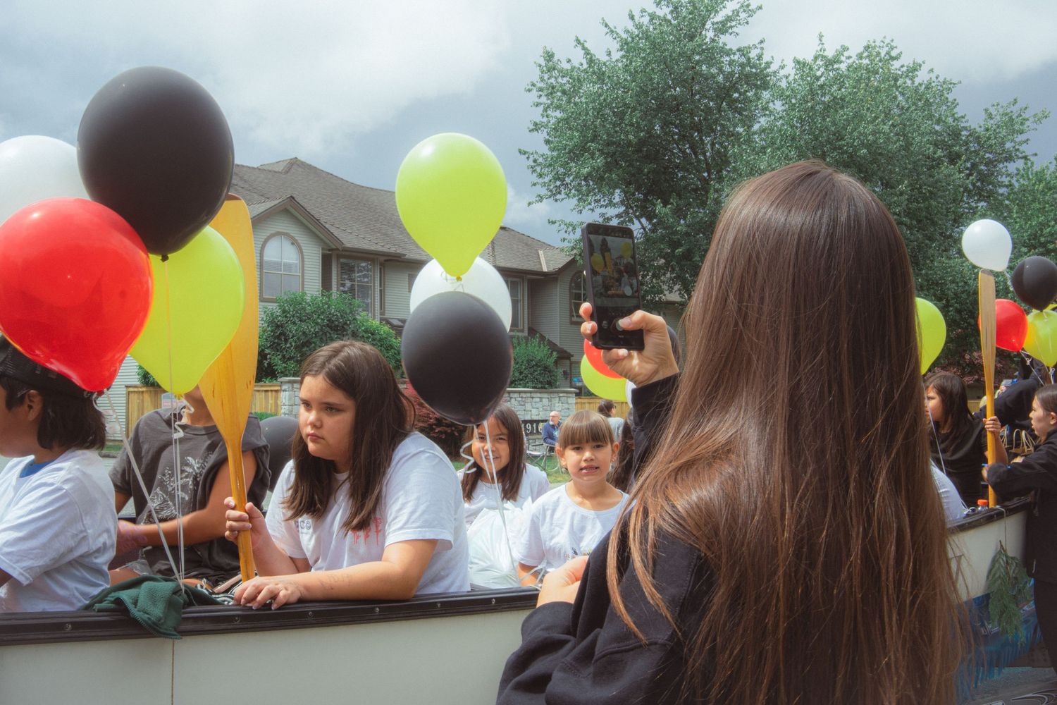 Children holding colorful balloons on a parade float while a girl photographs them from behind.