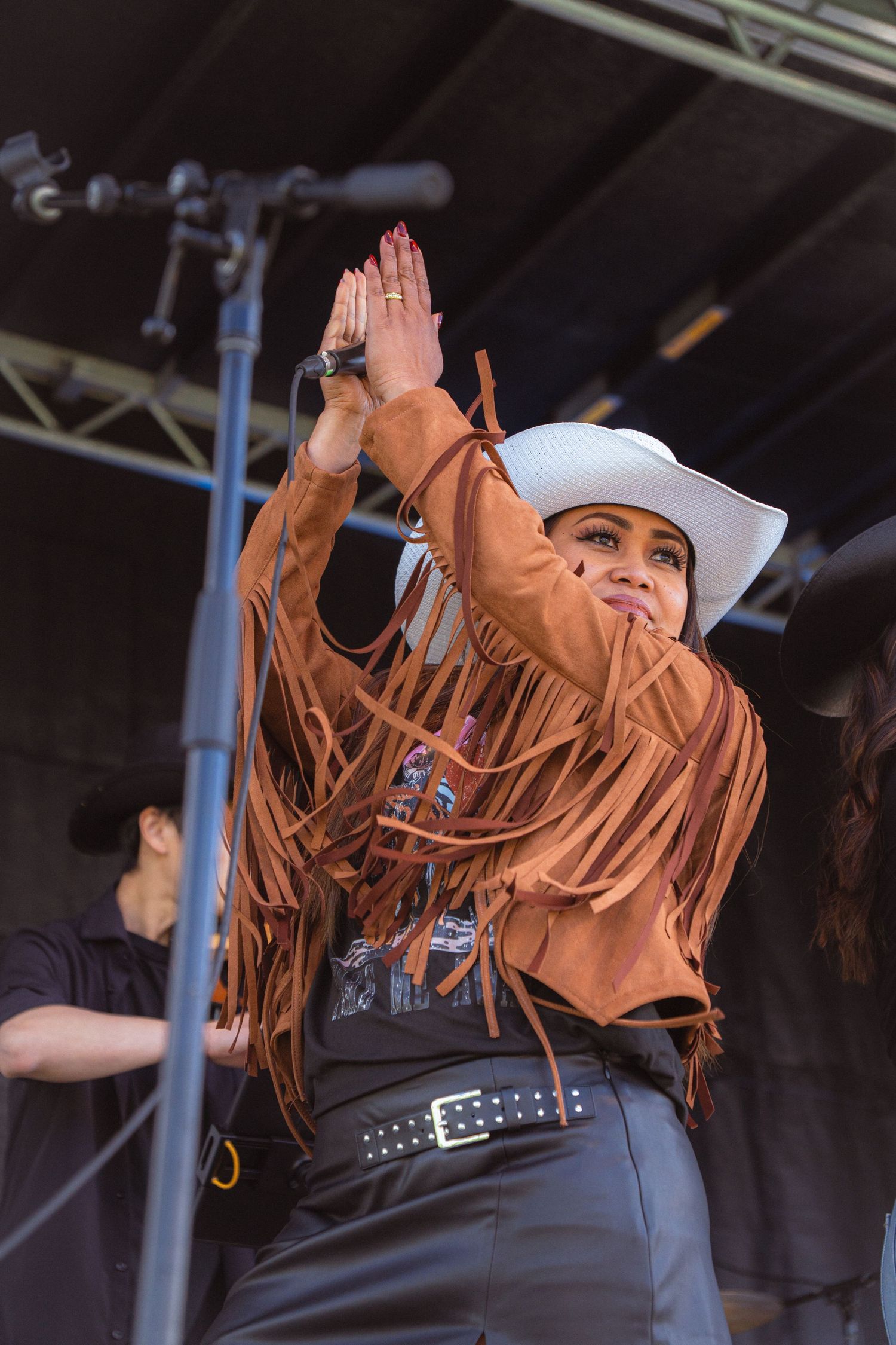 Woman in white cowboy hat and brown fringe jacket raises hands on stage at outdoor country music festival.
