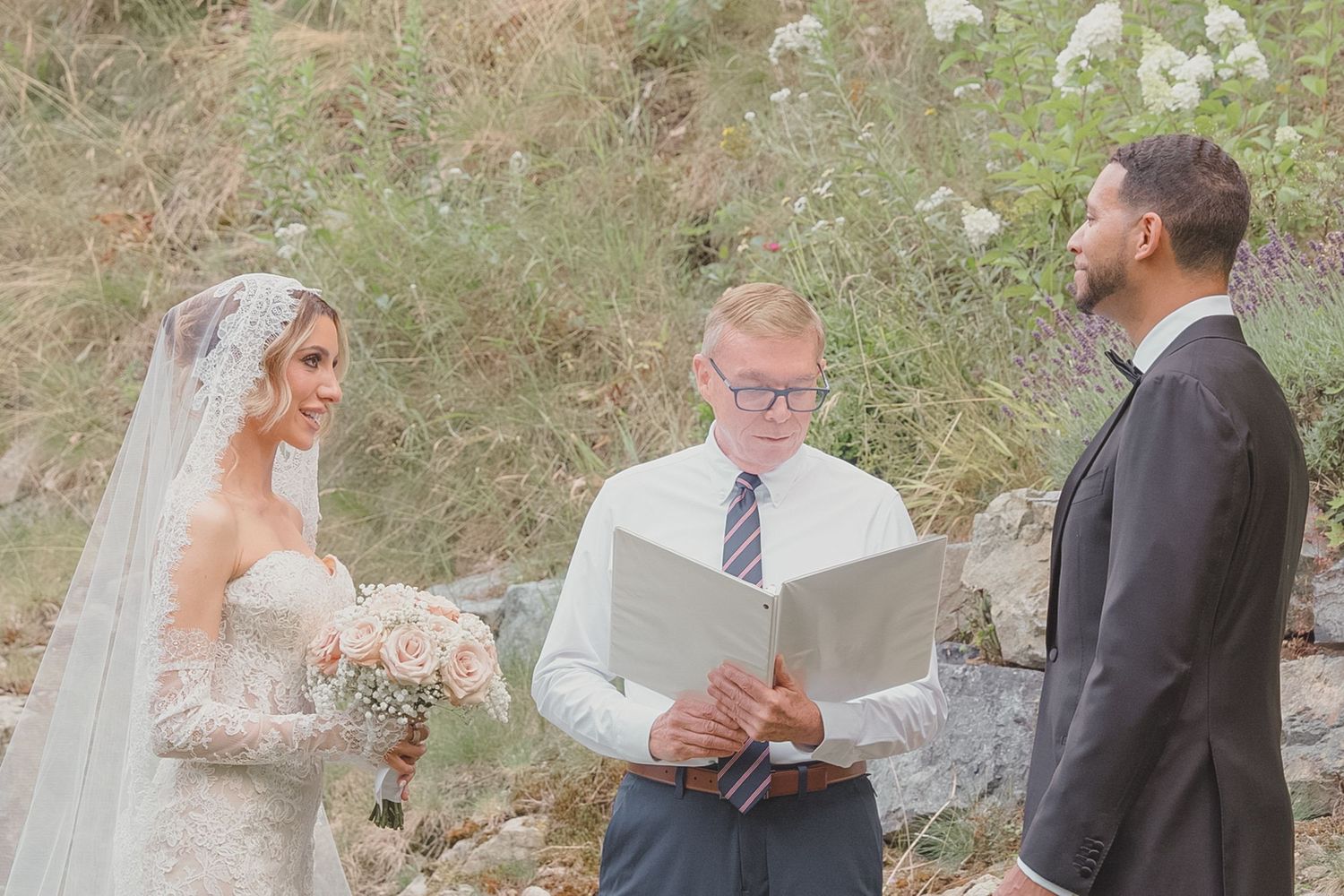 Bride and groom exchange vows outdoors with officiant reading from book during elegant wedding ceremony.