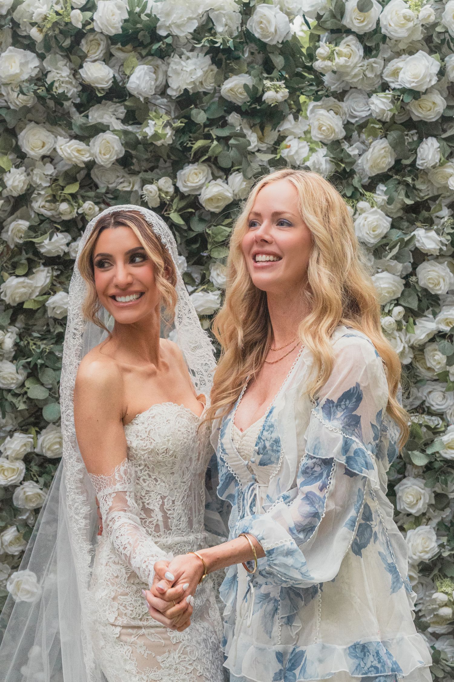Bride in lace gown and veil poses with blonde woman in floral dress before white rose flower wall backdrop.