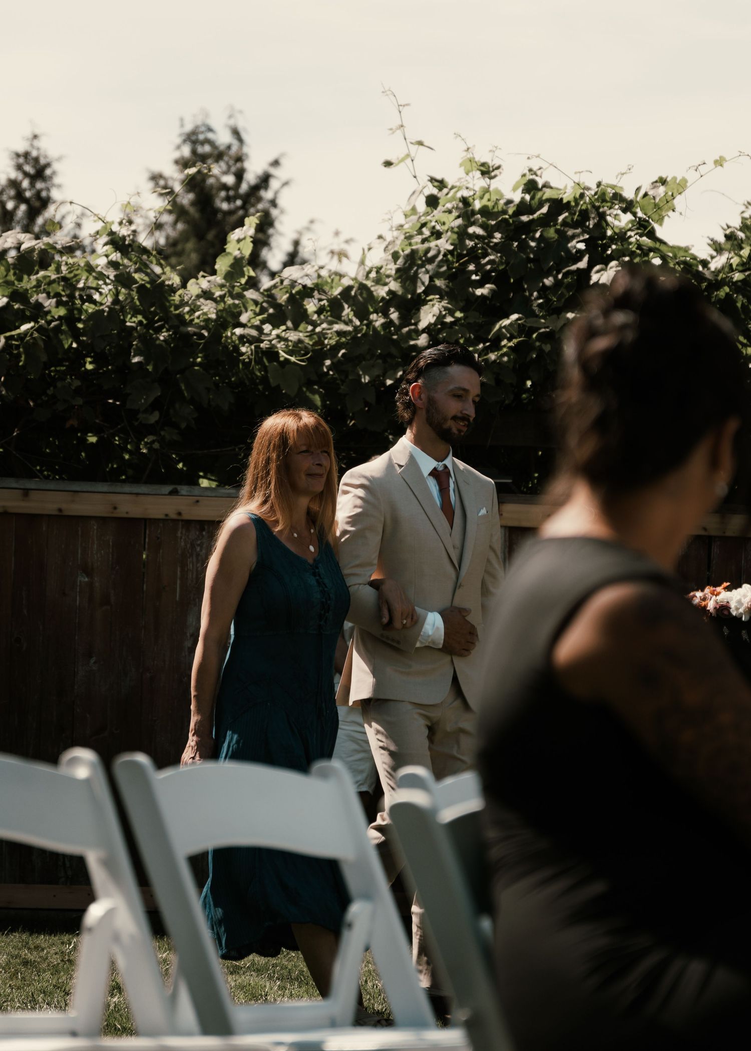 Groom in beige suit walks with woman in teal dress at outdoor wedding ceremony with white chairs.