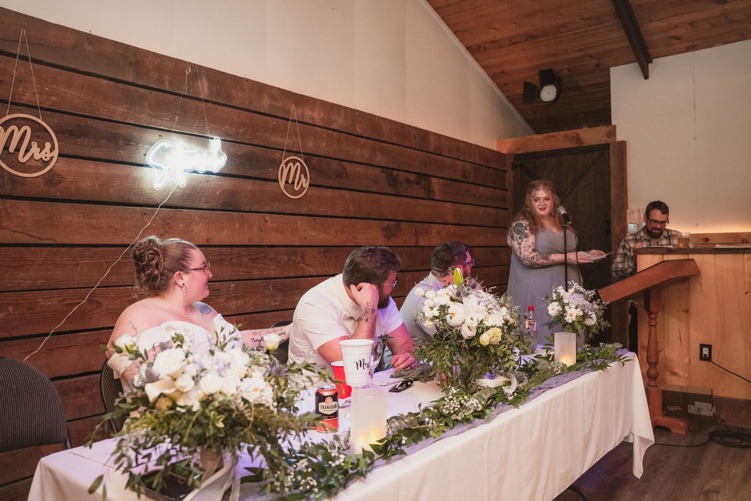 Wedding party seated at decorated head table with white floral arrangements while officiant speaks at rustic barn reception.
